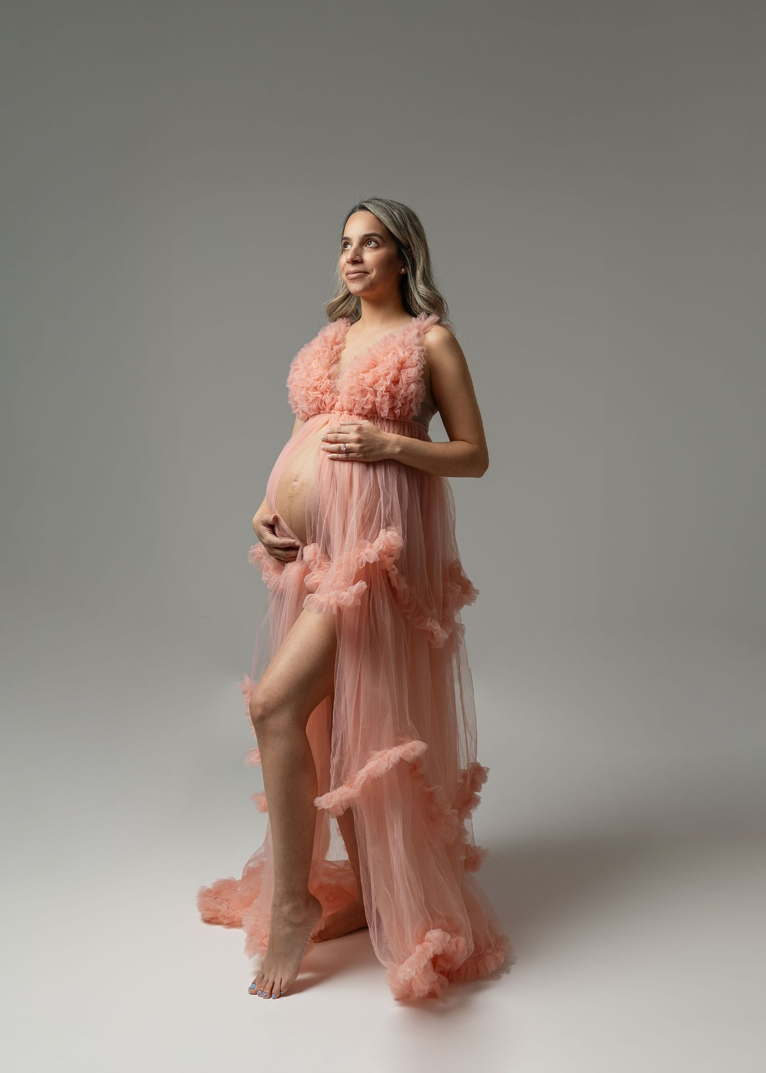 Maternity portrait in flowing coral gown against neutral backdrop.