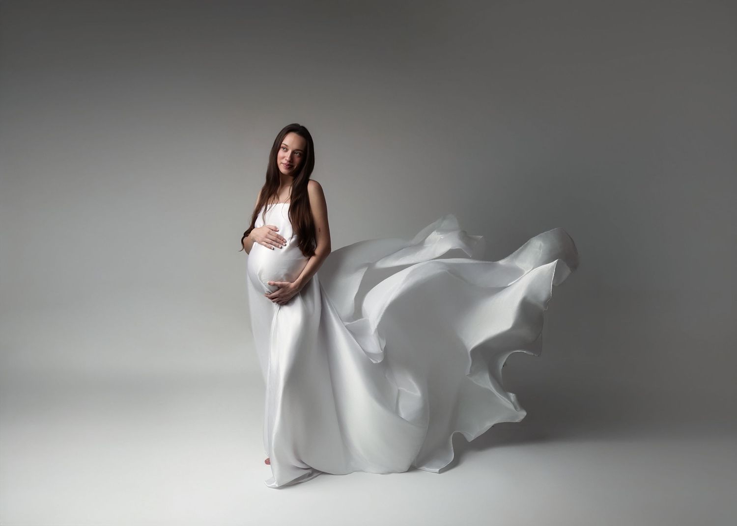 A flowing white maternity gown billows dramatically against a gray studio backdrop.