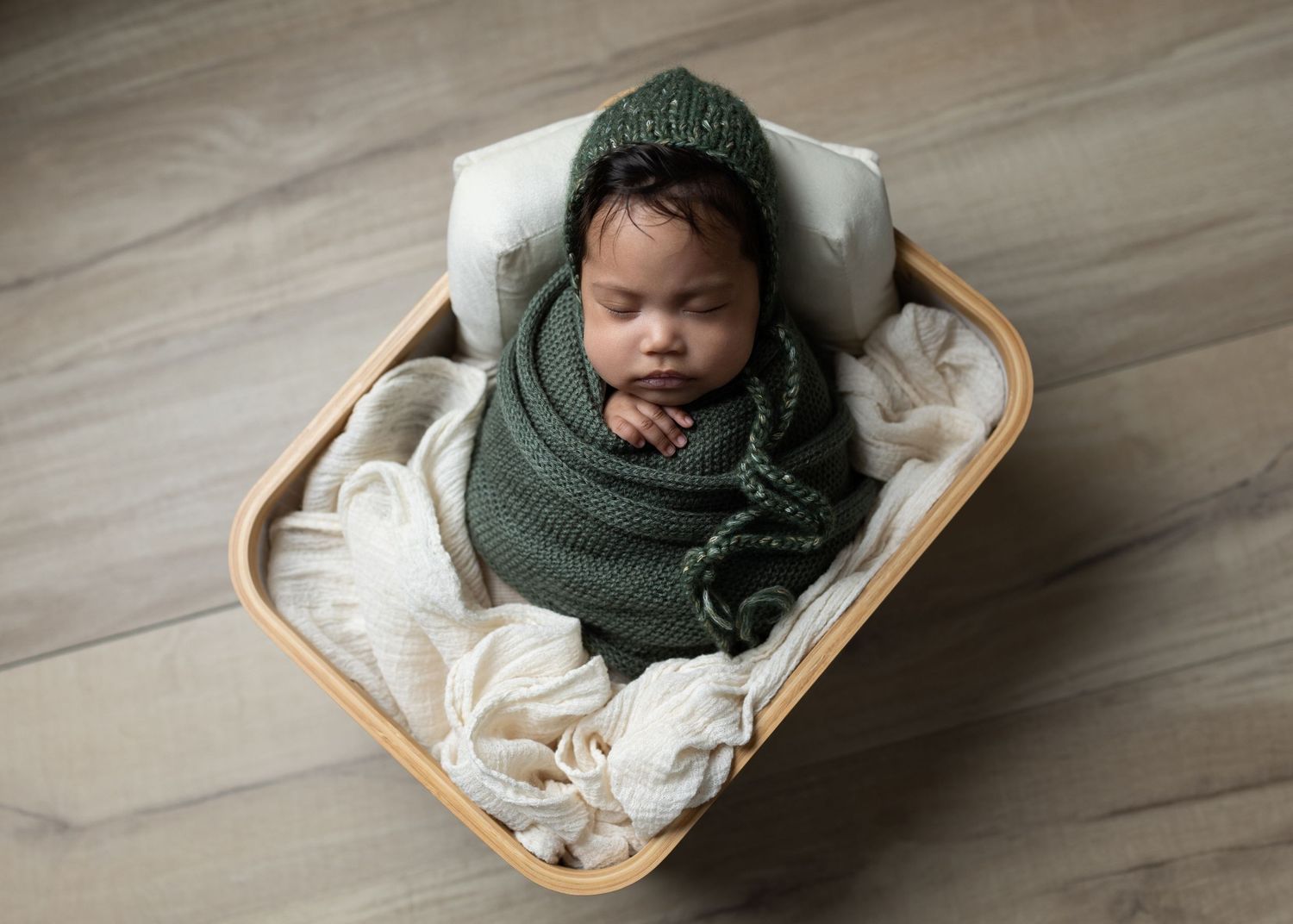 Newborn baby wrapped in green swaddle lying in wooden basket on hardwood floor.
