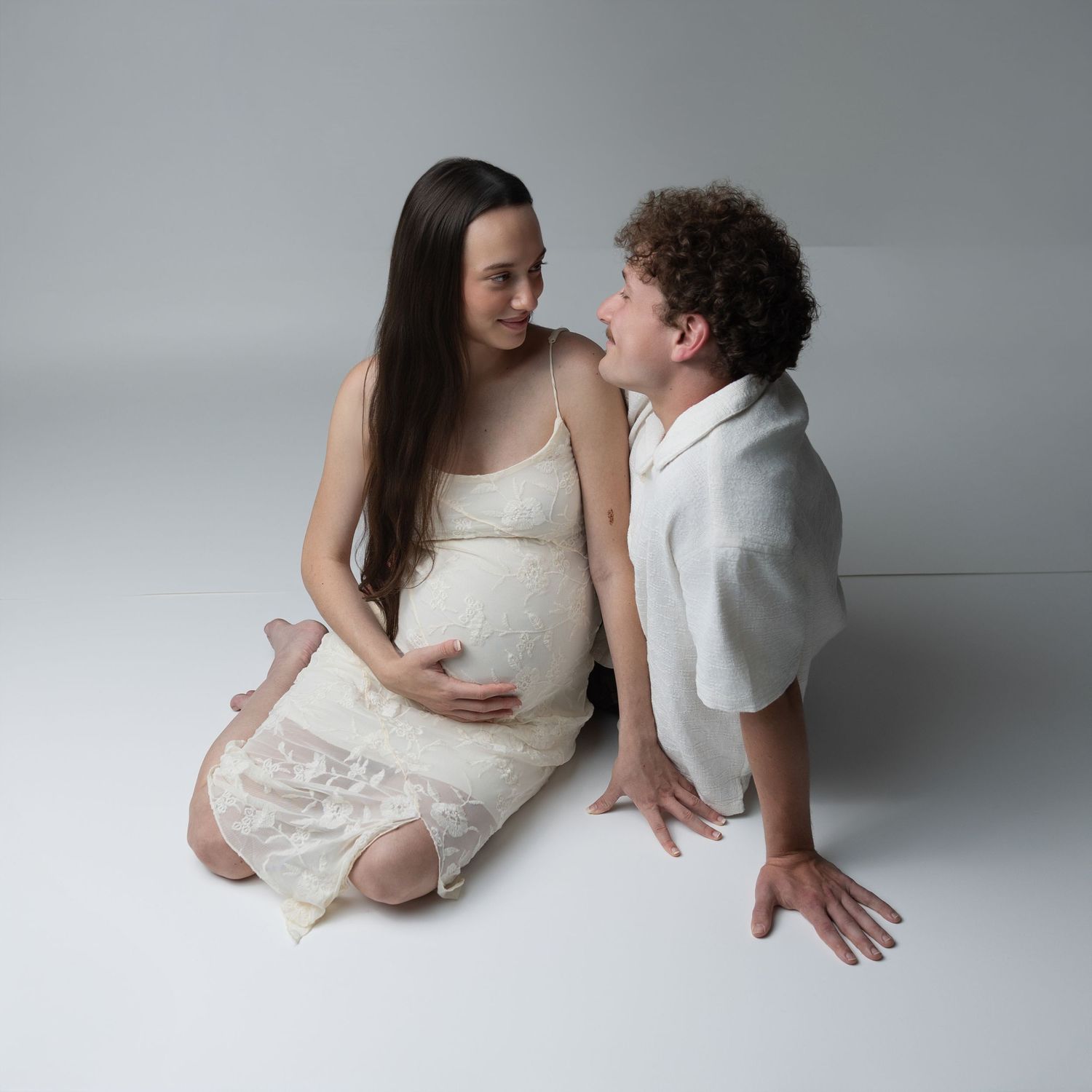 A series of intimate maternity photos showing a couple in white clothing against a light gray studio backdrop.