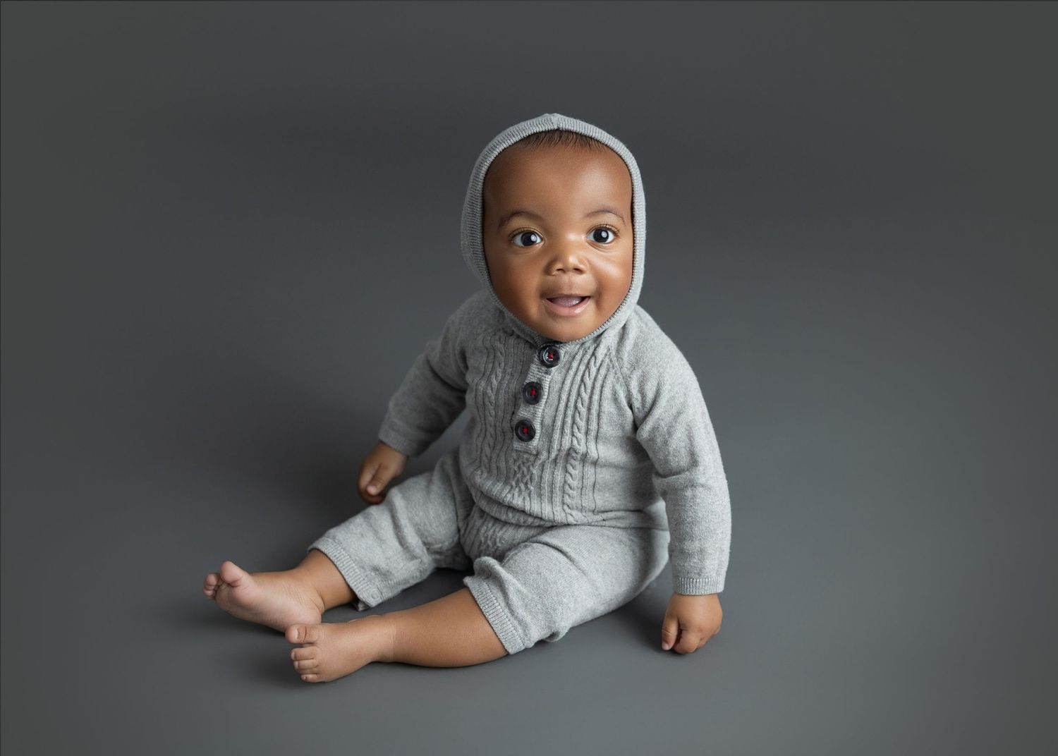 Baby wearing grey knit romper sitting on grey background with joyful expression.