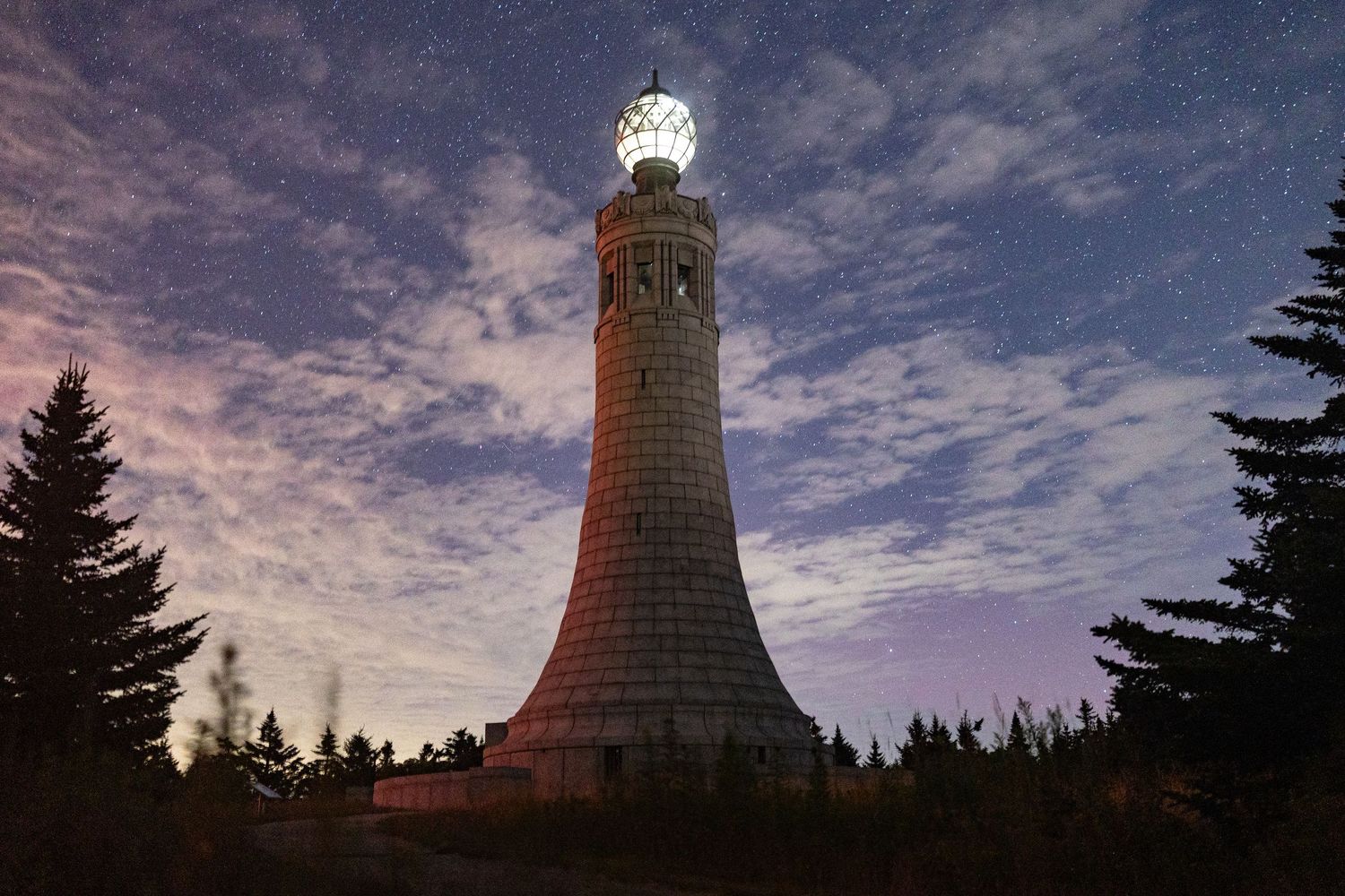 Dramatic sunset sky over a lighthouse with evergreen trees silhouetted in the foreground.