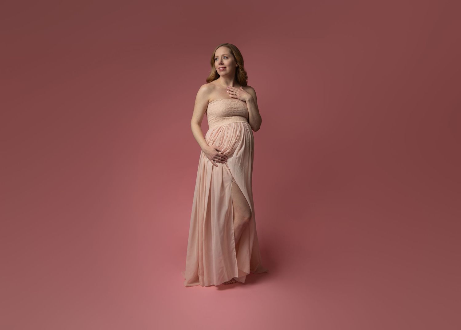 A pregnant woman in a flowing beige maternity dress poses elegantly against a pink studio backdrop.