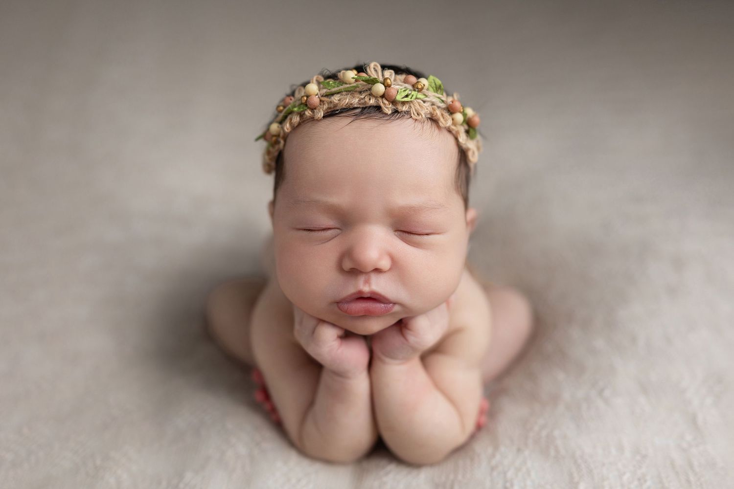 Sleeping newborn baby wearing a delicate floral headband, posed with hands under chin on a soft white blanket.