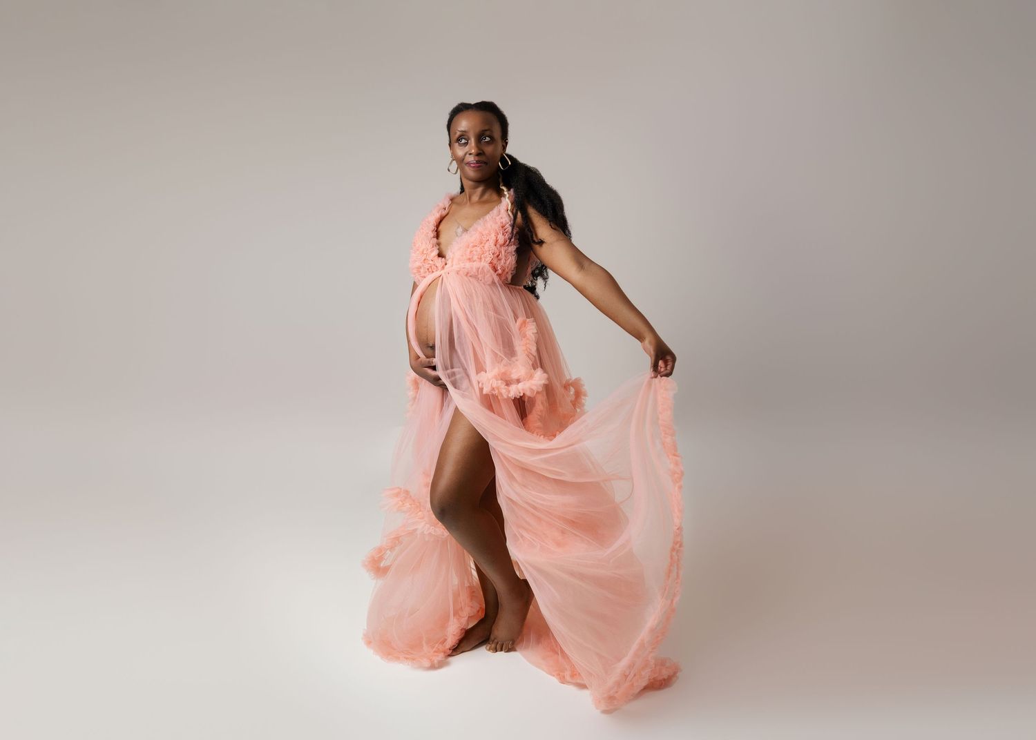 Maternity photo session featuring flowing pink dress against light gray backdrop.