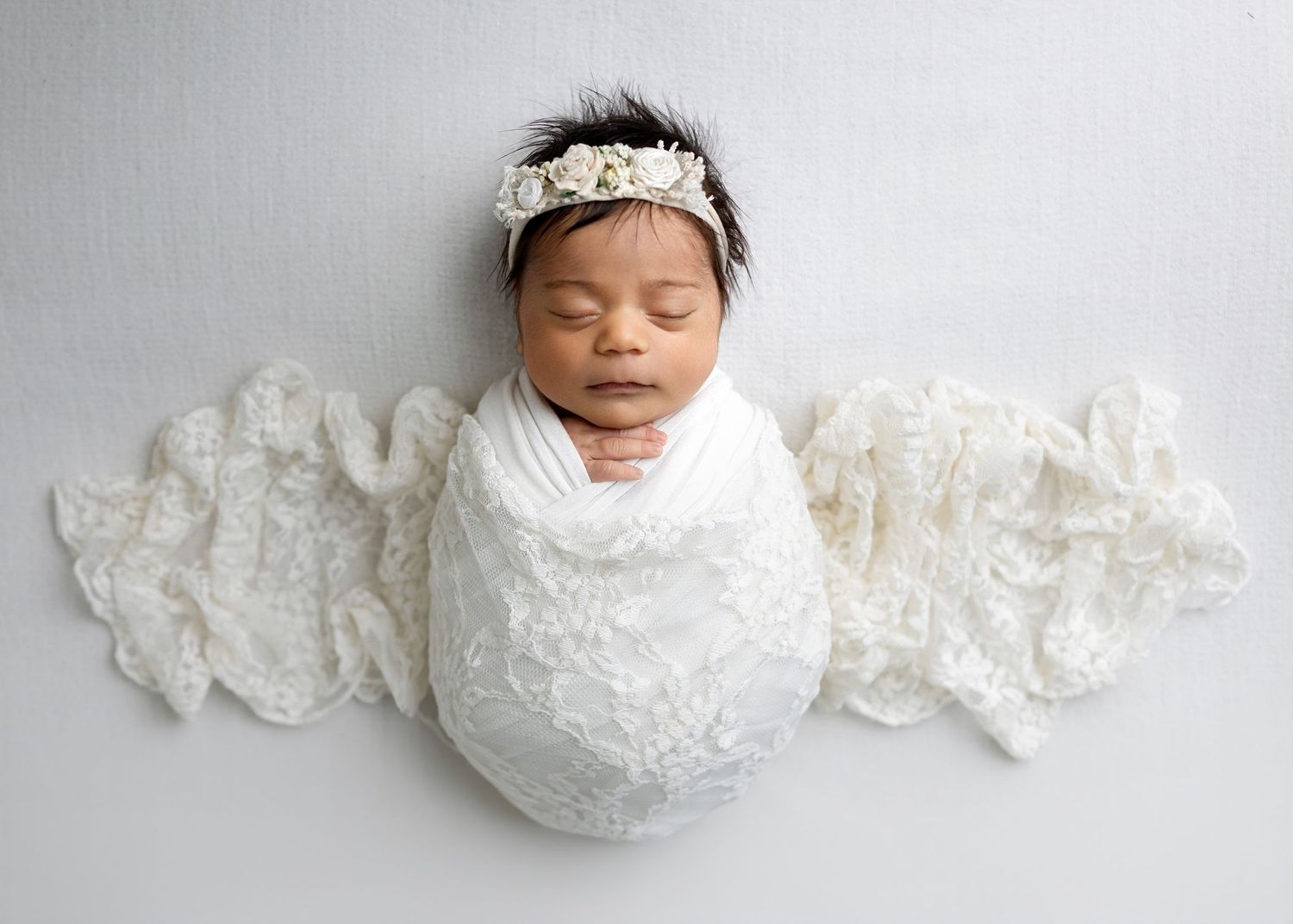 Sleeping newborn baby swaddled in white lace wrap with floral headband on gray background.