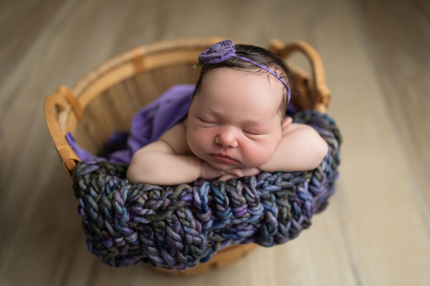 Newborn baby sleeping peacefully in a purple knitted wrap on a wooden backdrop.