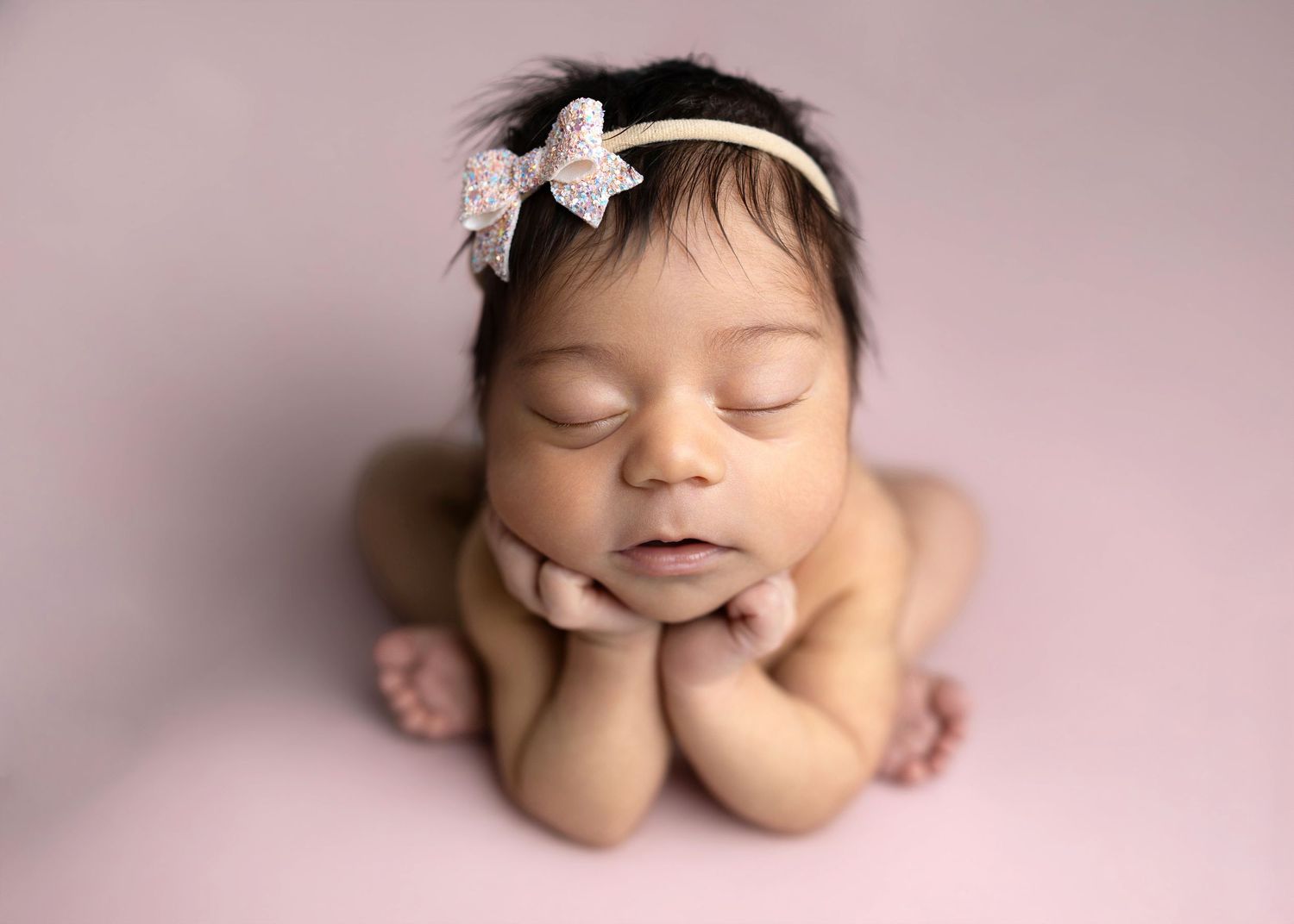 Newborn baby girl sleeping peacefully in froggy pose wearing a delicate bow headband on a soft pink background.