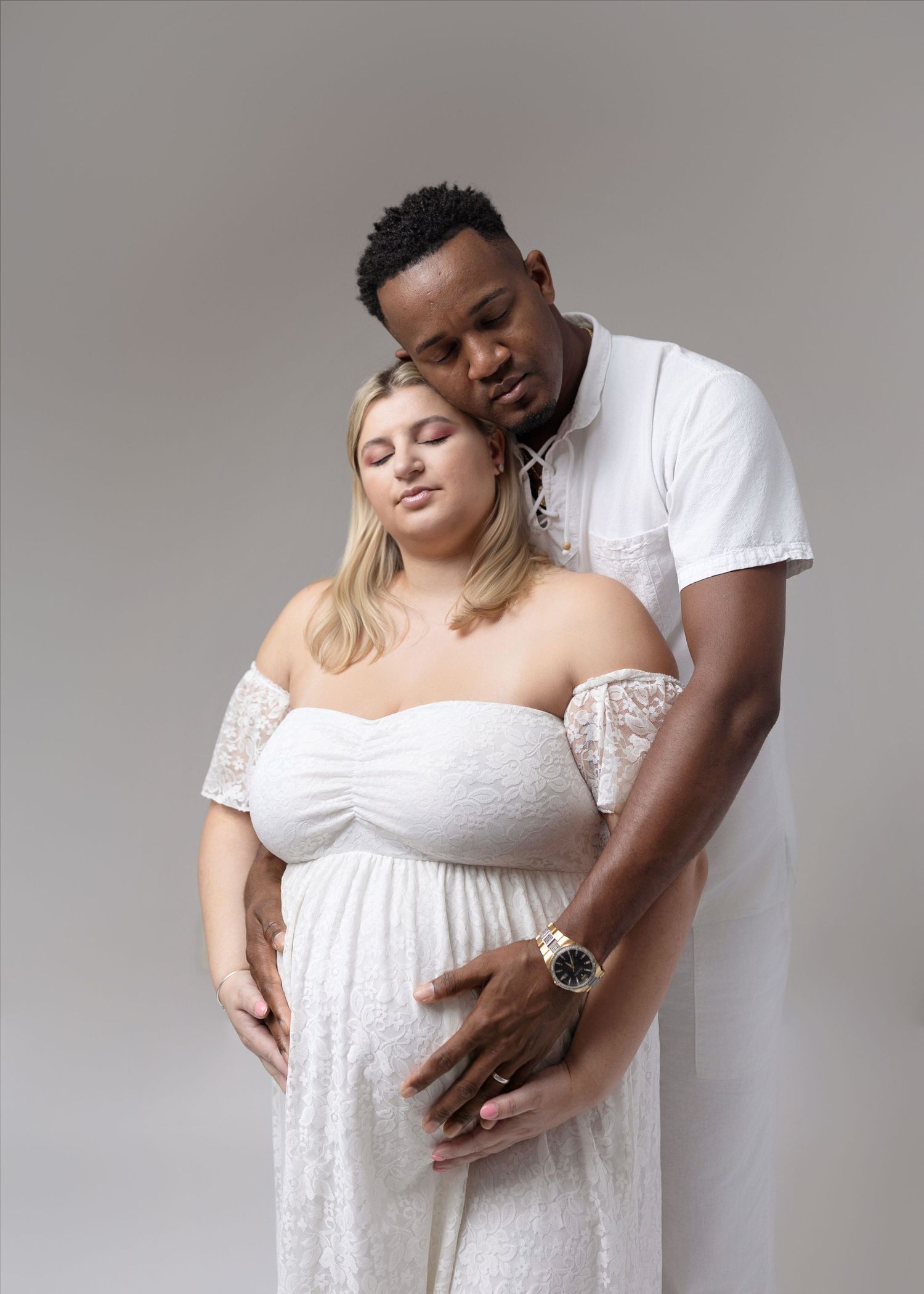 Intimate maternity poses of couple in white attire against neutral backdrop.
