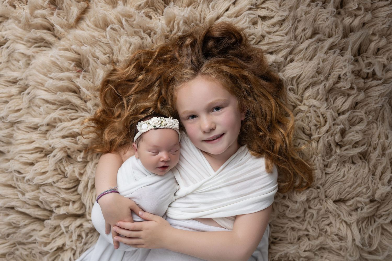 Two children cuddling on plush beige carpet in white outfits.