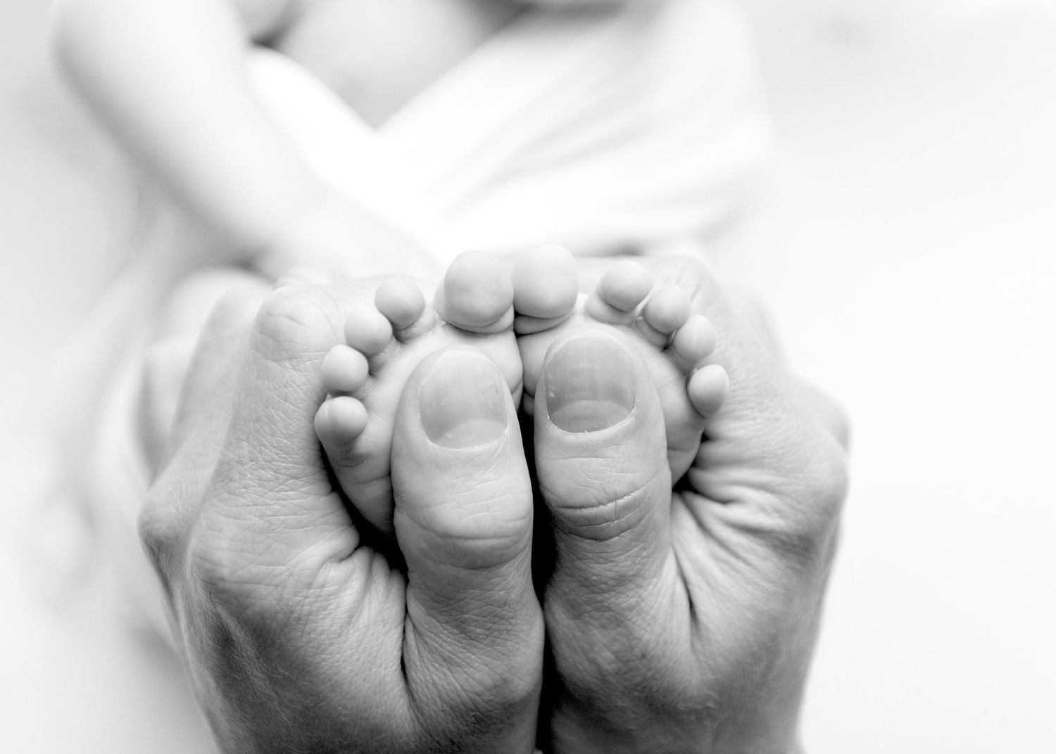 Adult hands gently cradling tiny newborn baby feet in a tender black and white close-up photo.