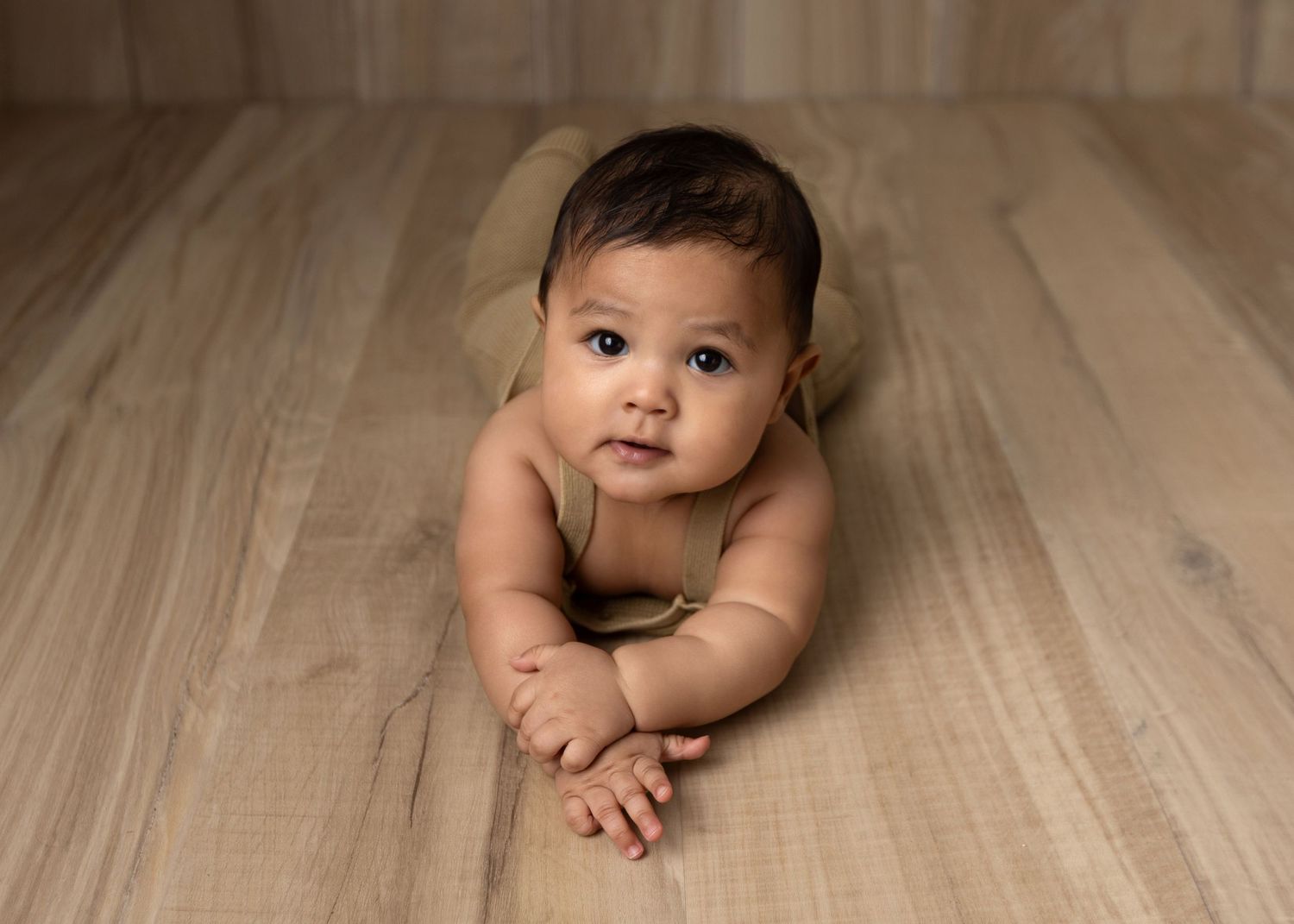 Adorable baby doing tummy time on a light wood floor, looking up with curious eyes, wearing a neutral wrap.