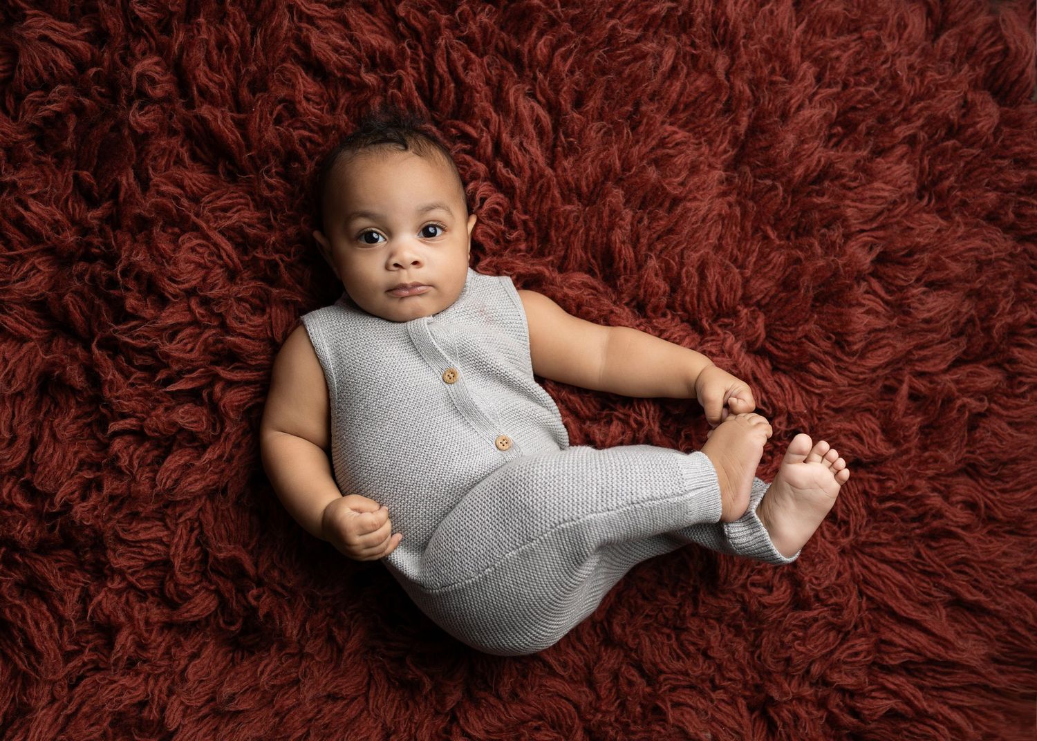 Baby wearing gray onesie photographed in various poses on burgundy textured rug.
