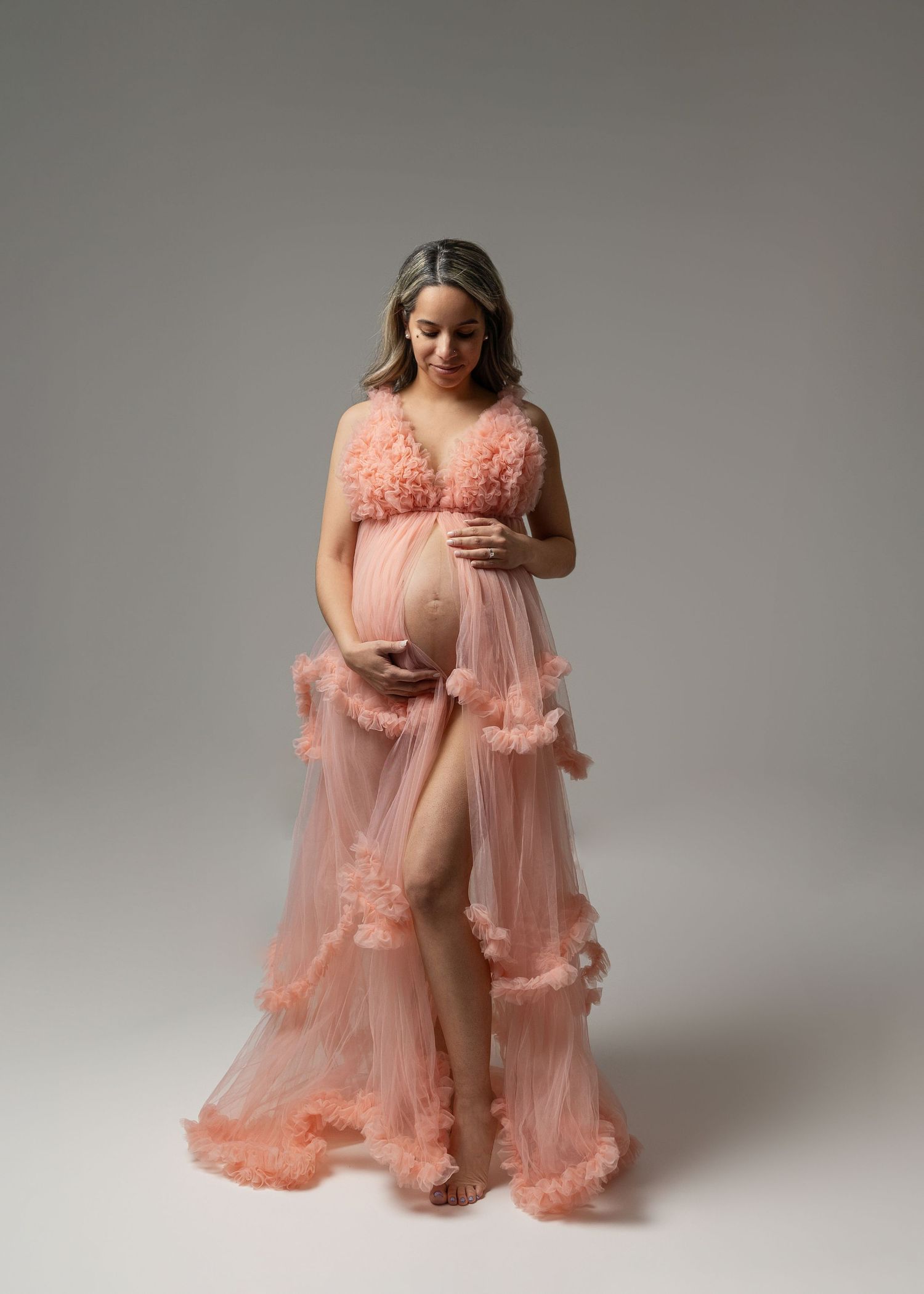 Romantic maternity photoshoot featuring flowing peach tulle gown against grey backdrop.
