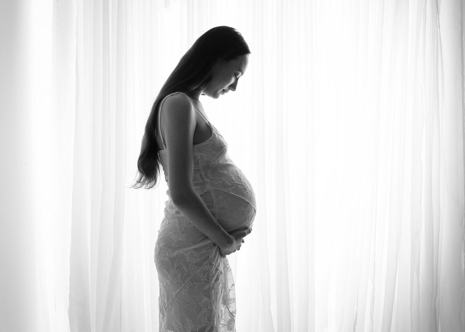 Black and white silhouette of a pregnant woman in lace dress cradling her baby bump by a bright window.