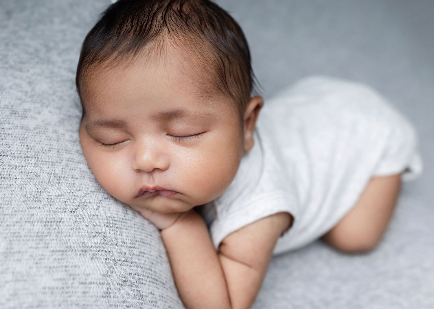 A sleeping newborn baby in a white onesie lies peacefully on soft gray fabric in a series of gentle poses.