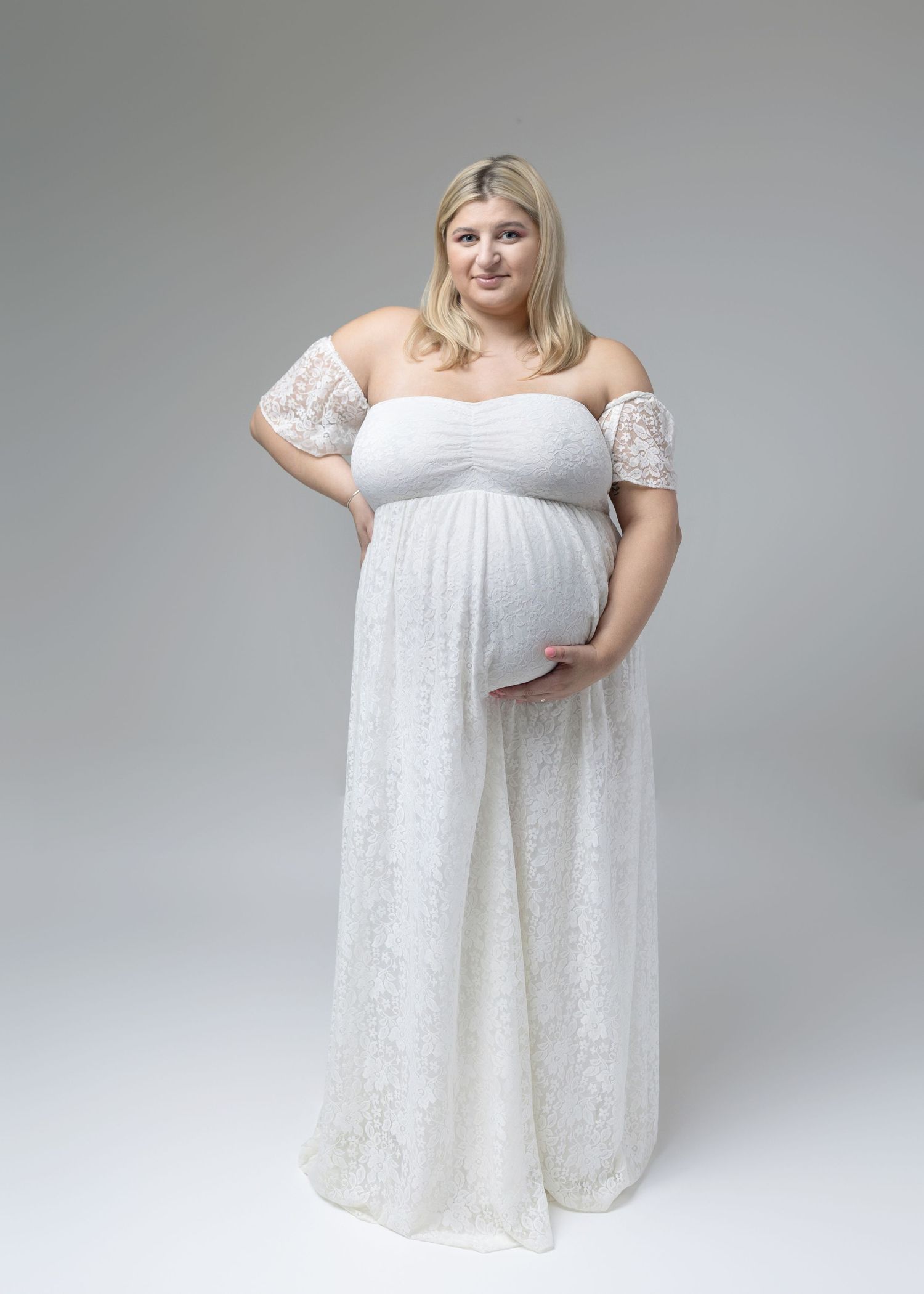 Elegant maternity portraits in white lace off-shoulder dress against grey background.
