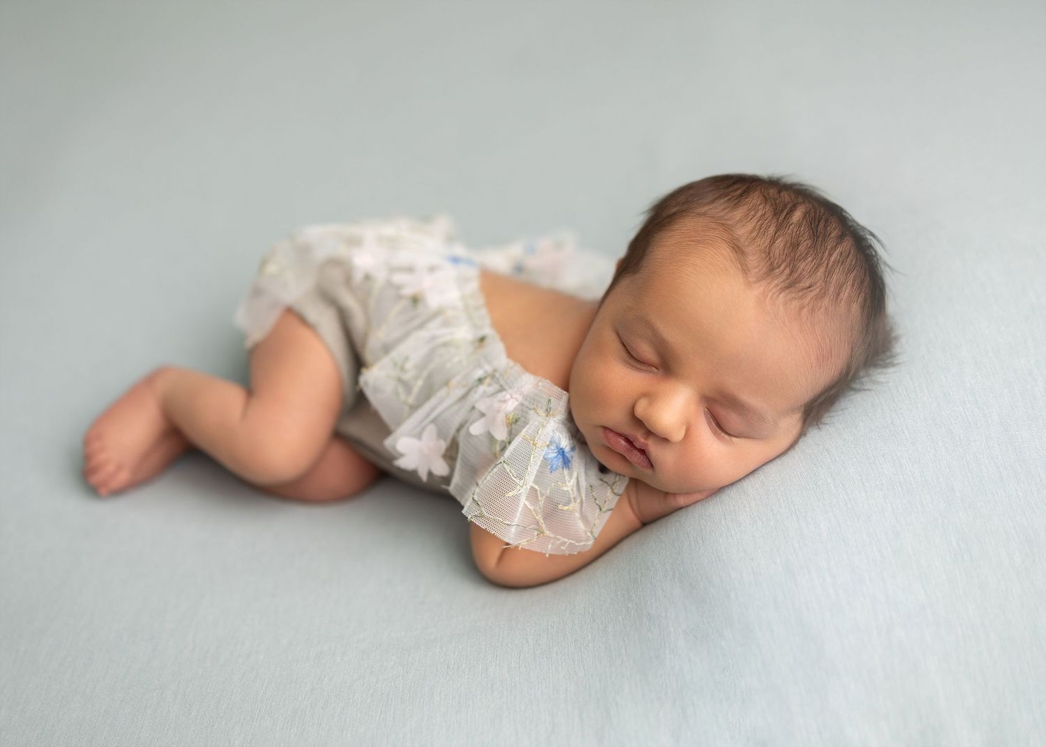 Peaceful newborn baby sleeping on a soft blue blanket wearing a delicate white romper in a professional photo.