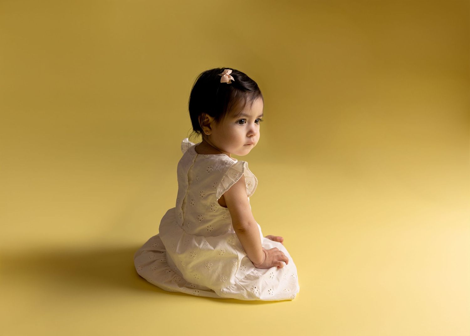 Baby in white dress poses against warm golden backdrop during studio session.