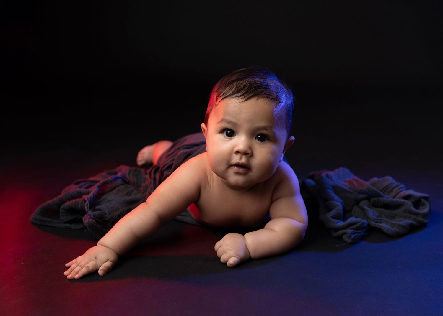 Adorable baby doing tummy time on dark fabric with dramatic blue and red studio lighting on black background.