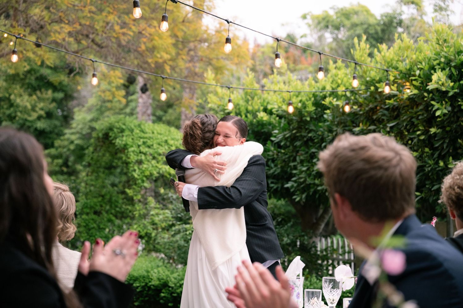Natural editorial documentary moment of a couple embracing at an outdoor celebration with string lights, greenery, and guests clapping.