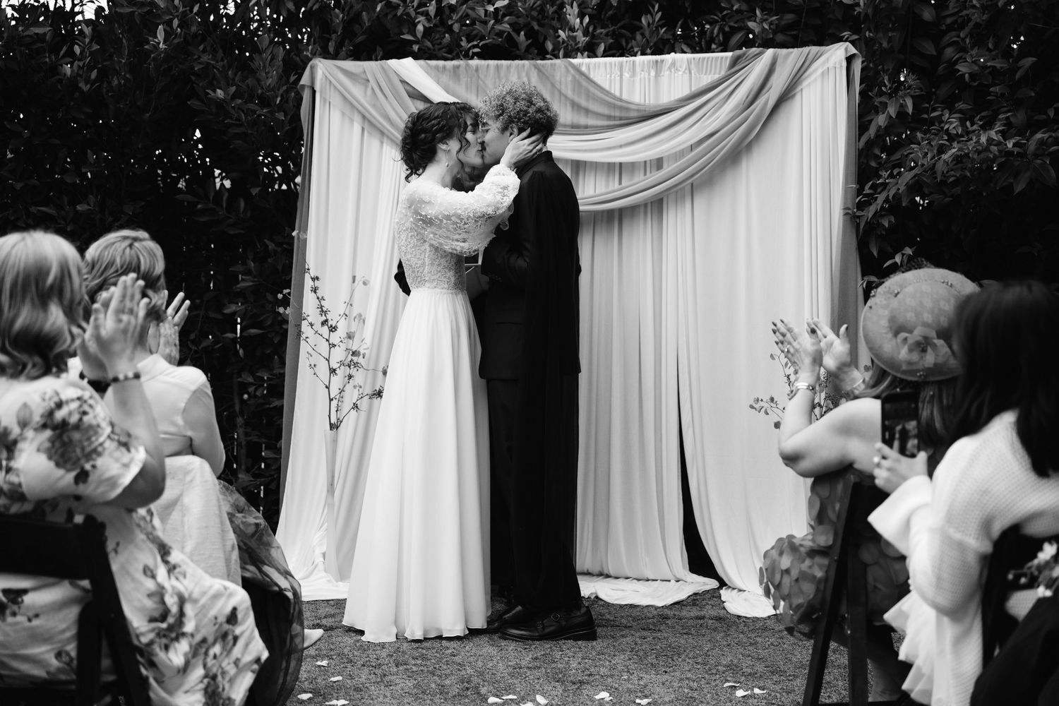 Romantic editorial documentary wedding moment of a couple kissing under a draped arch as guests watch and applaud in black and white.