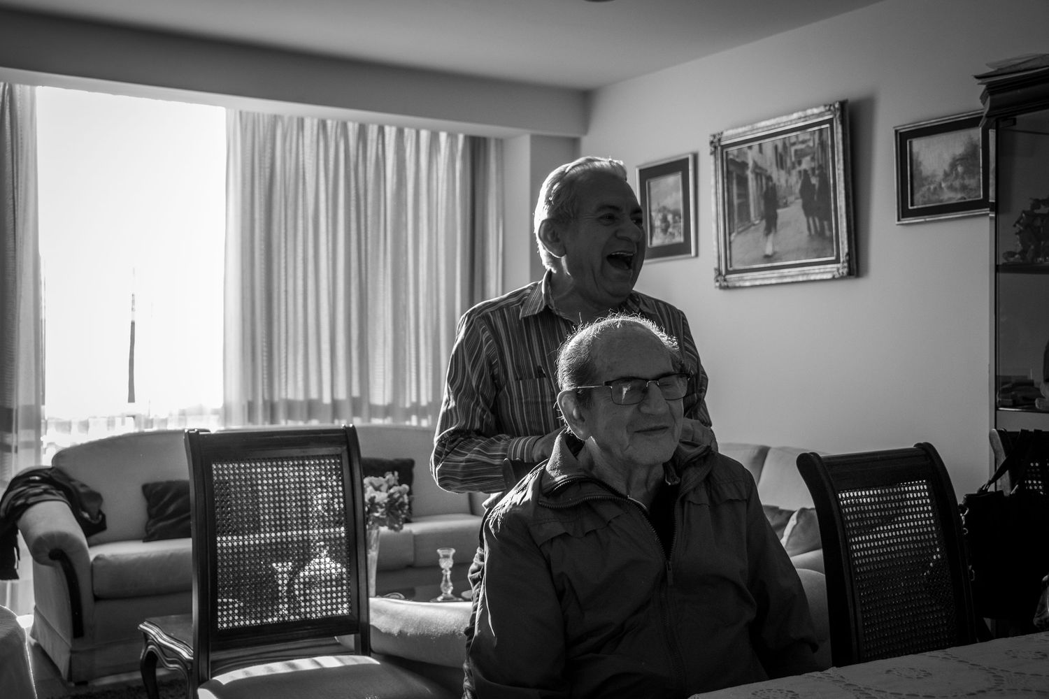 Editorial documentary black‑and‑white moment of two older adults sharing conversation in a sunlit living room with warm, nostalgic light.