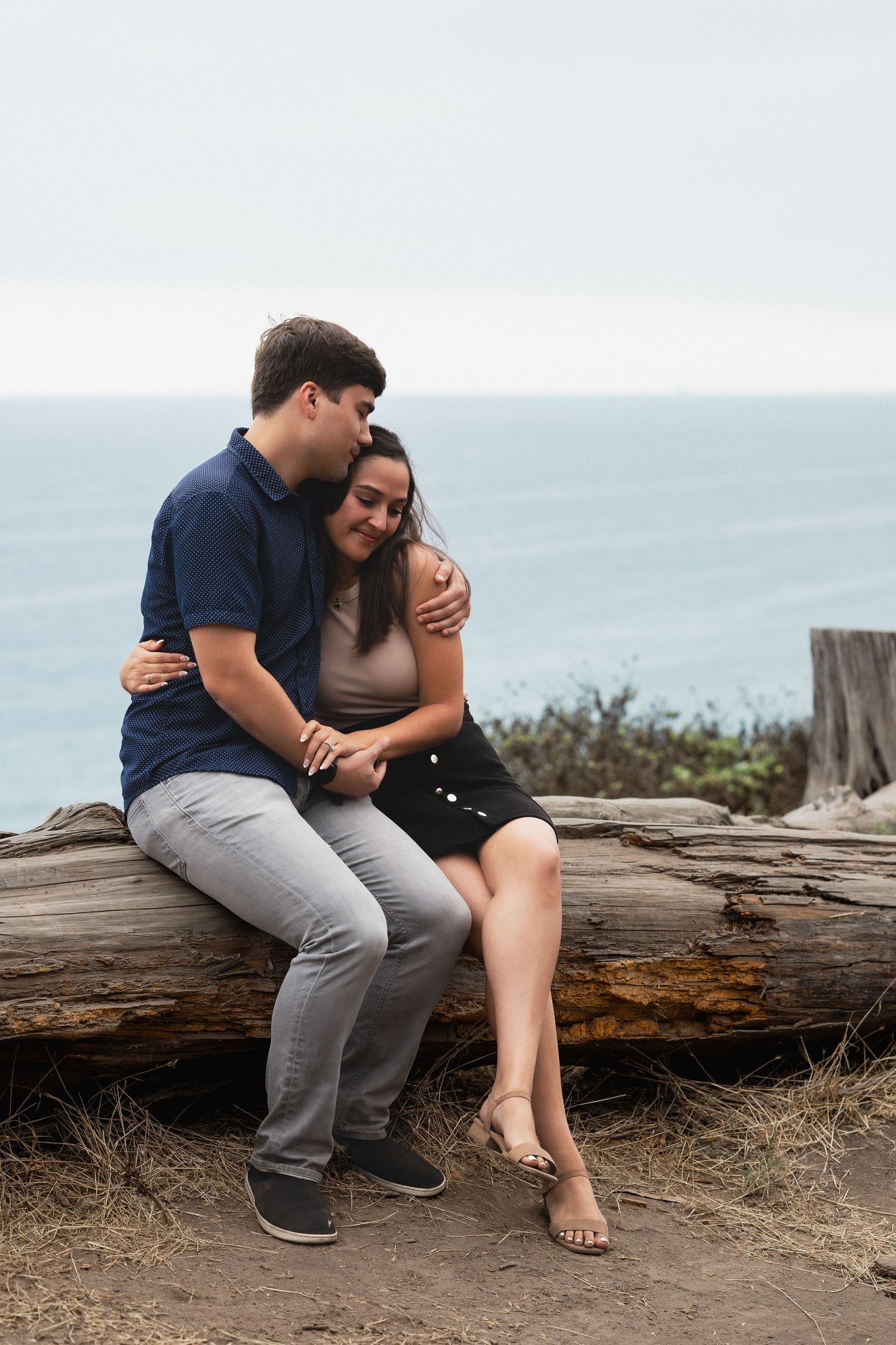 Natural editorial documentary moment of a couple sitting on driftwood by the coast, sharing a quiet embrace with ocean and clouds behind the
