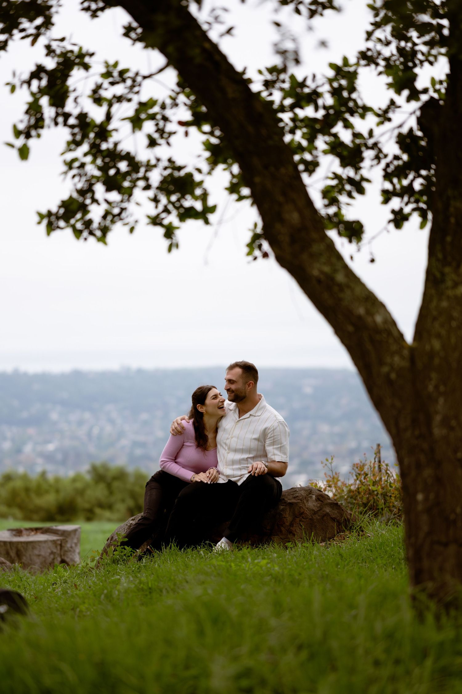 Couple laughing on a rock beneath an oak tree at Franceschi Park Santa Barbara, engagement photography, Herics Photo