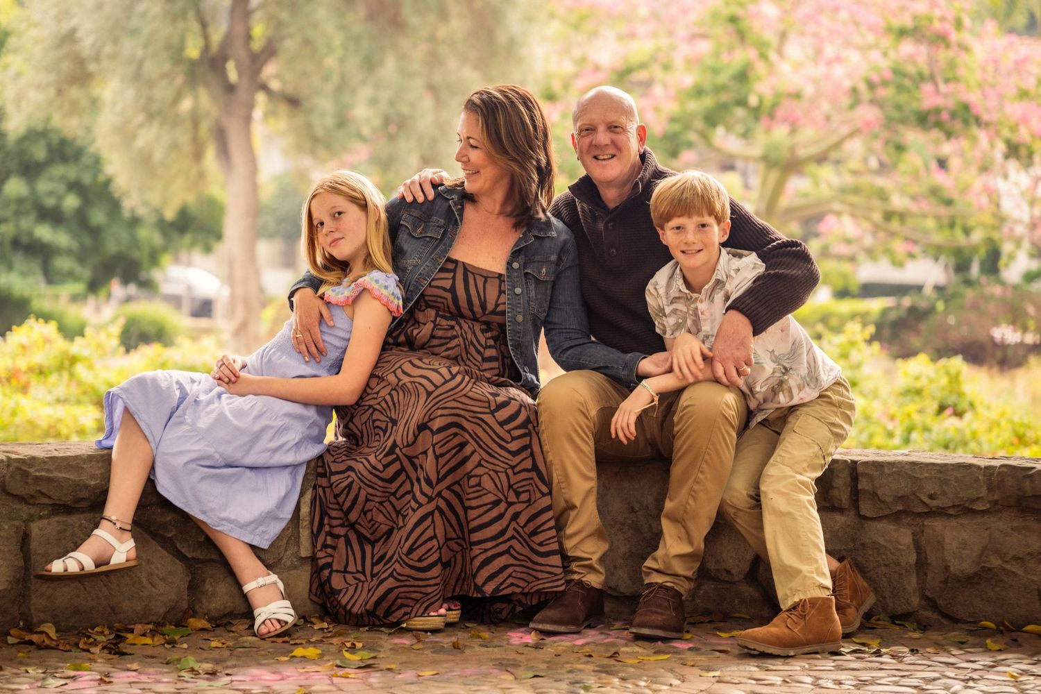 Editorial documentary family photo of two parents and two kids sitting on a stone ledge surrounded by greenery and soft natural light.