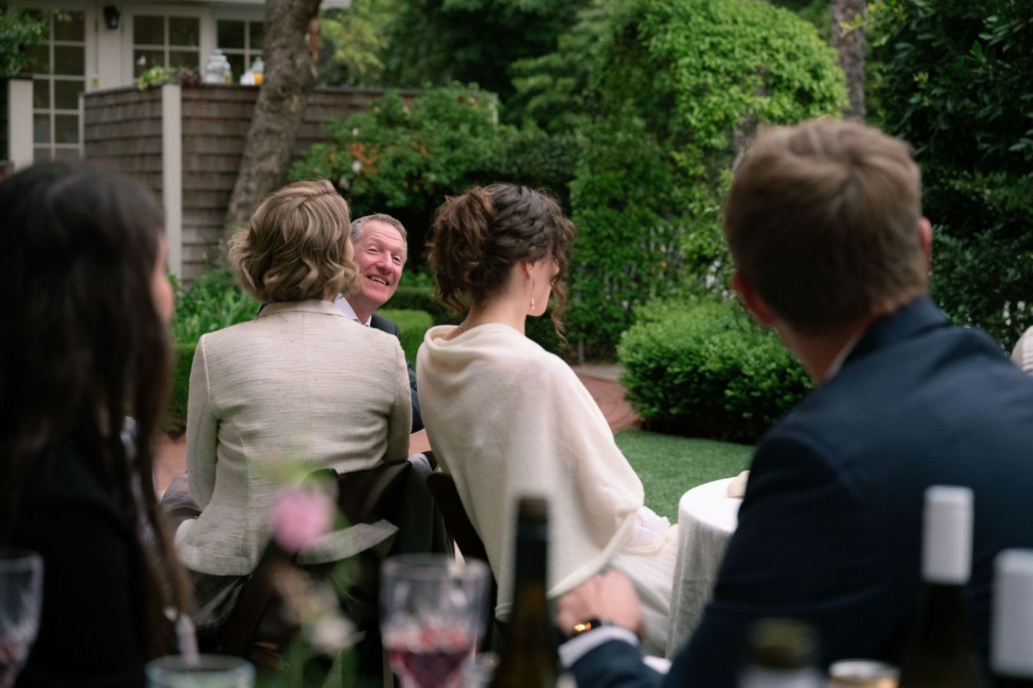 Natural editorial documentary moment of wedding guests talking at an outdoor reception table surrounded by greenery and soft light.