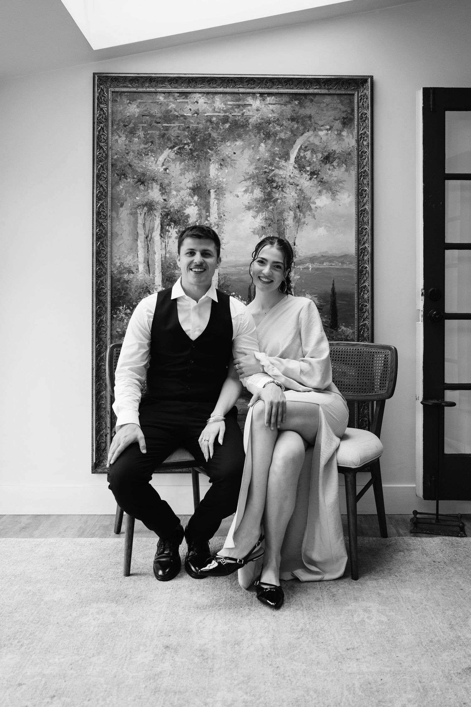 Natural editorial documentary wedding moment of a couple sitting indoors beneath a large landscape painting in soft black‑and‑white light.