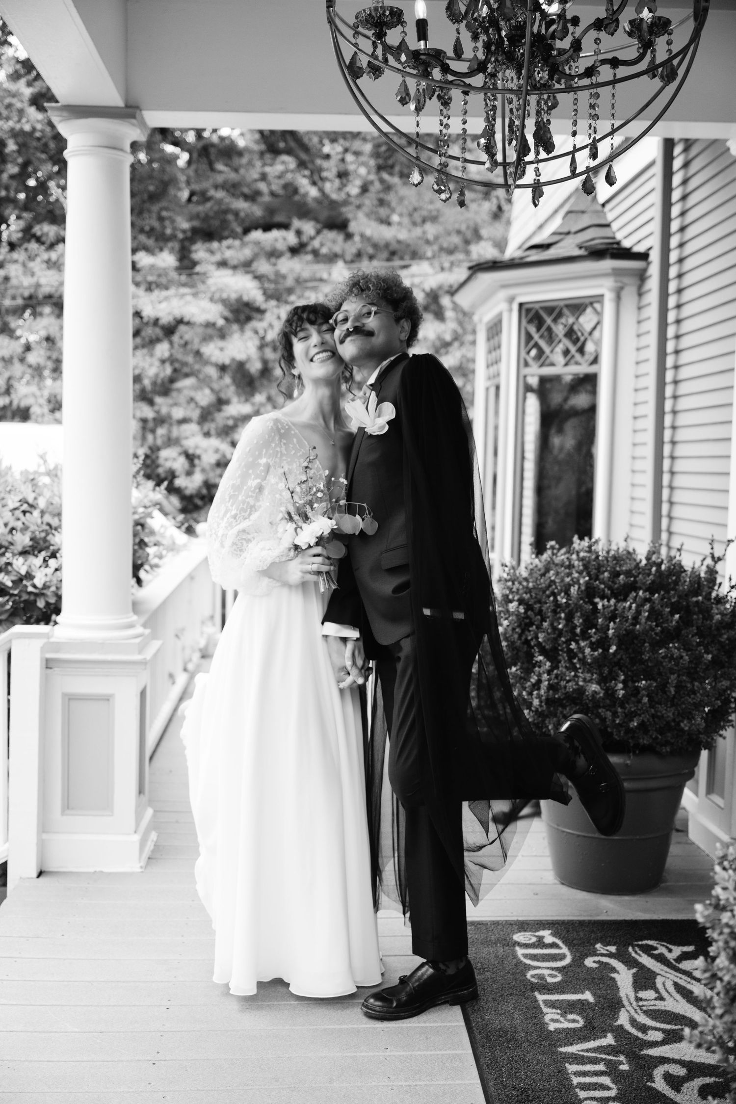 Natural editorial documentary wedding moment of a couple on a porch, one lifting a leg playfully while holding flowers under a chandelier.