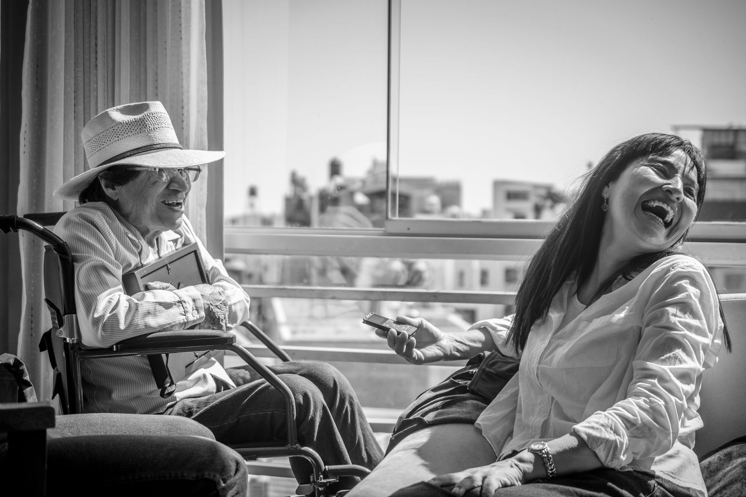 Soft editorial documentary scene of two people laughing near a bright window, one in a wheelchair, framed by daylight and city buildings.