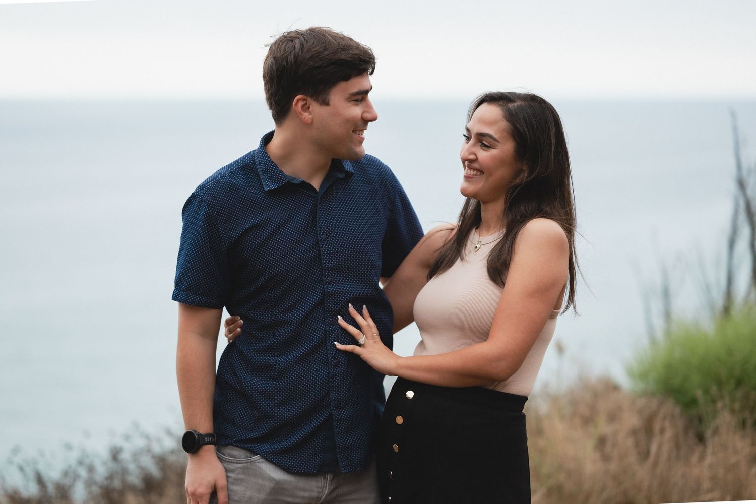 Natural editorial documentary proposal moment of a couple standing close by the water, sharing an intimate touch with soft coastal light.