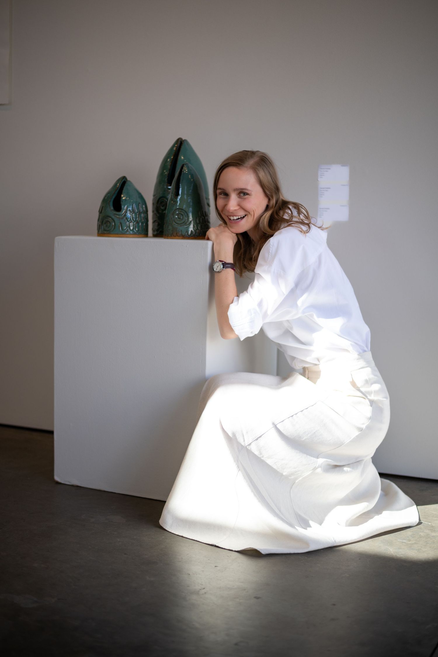Editorial documentary photo of a person kneeling beside a pedestal, observing two textured ceramic sculptures in soft studio light.