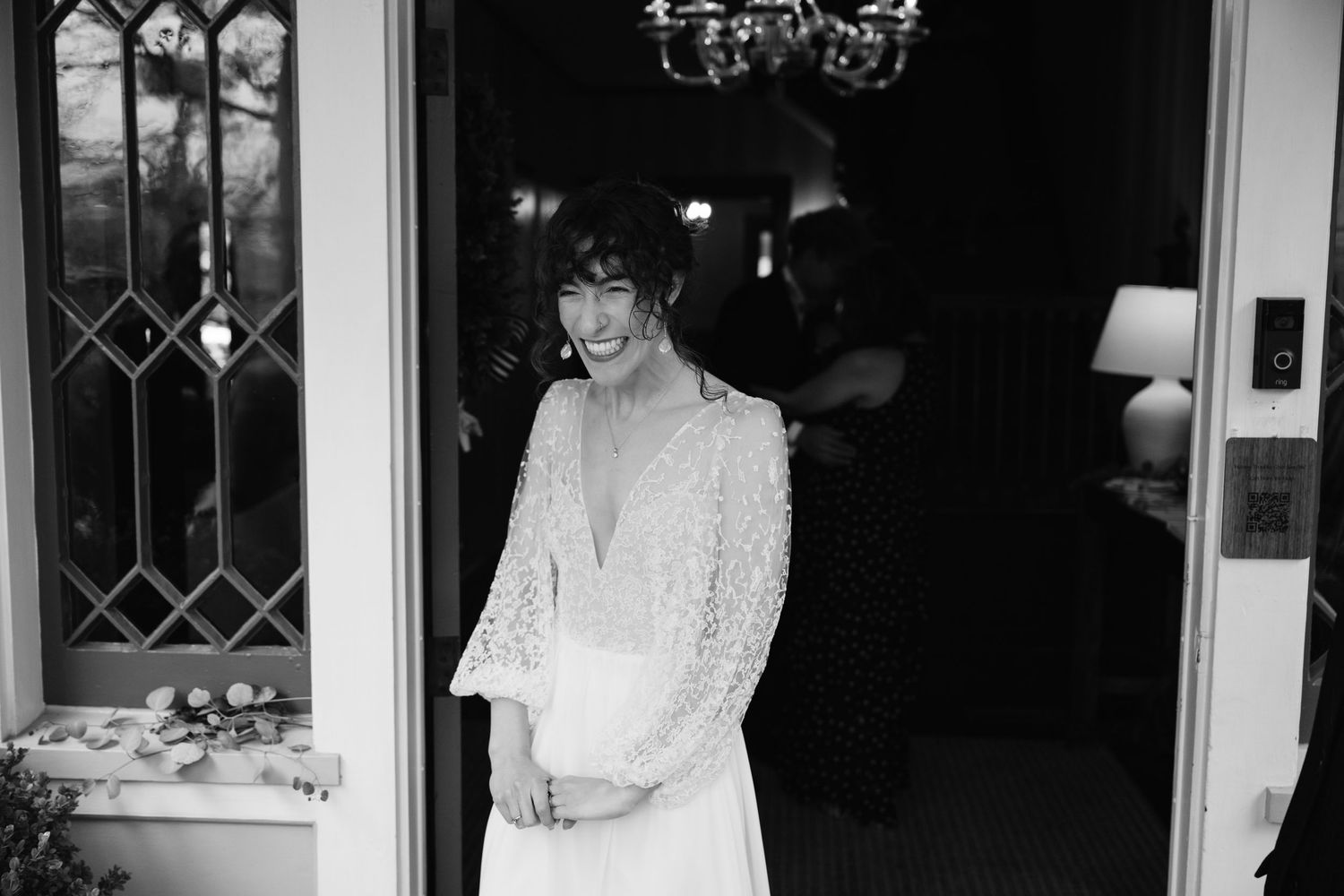 Natural editorial documentary wedding moment of a bride standing in a doorway with soft light, flowers, and classic black‑and‑white tones.
