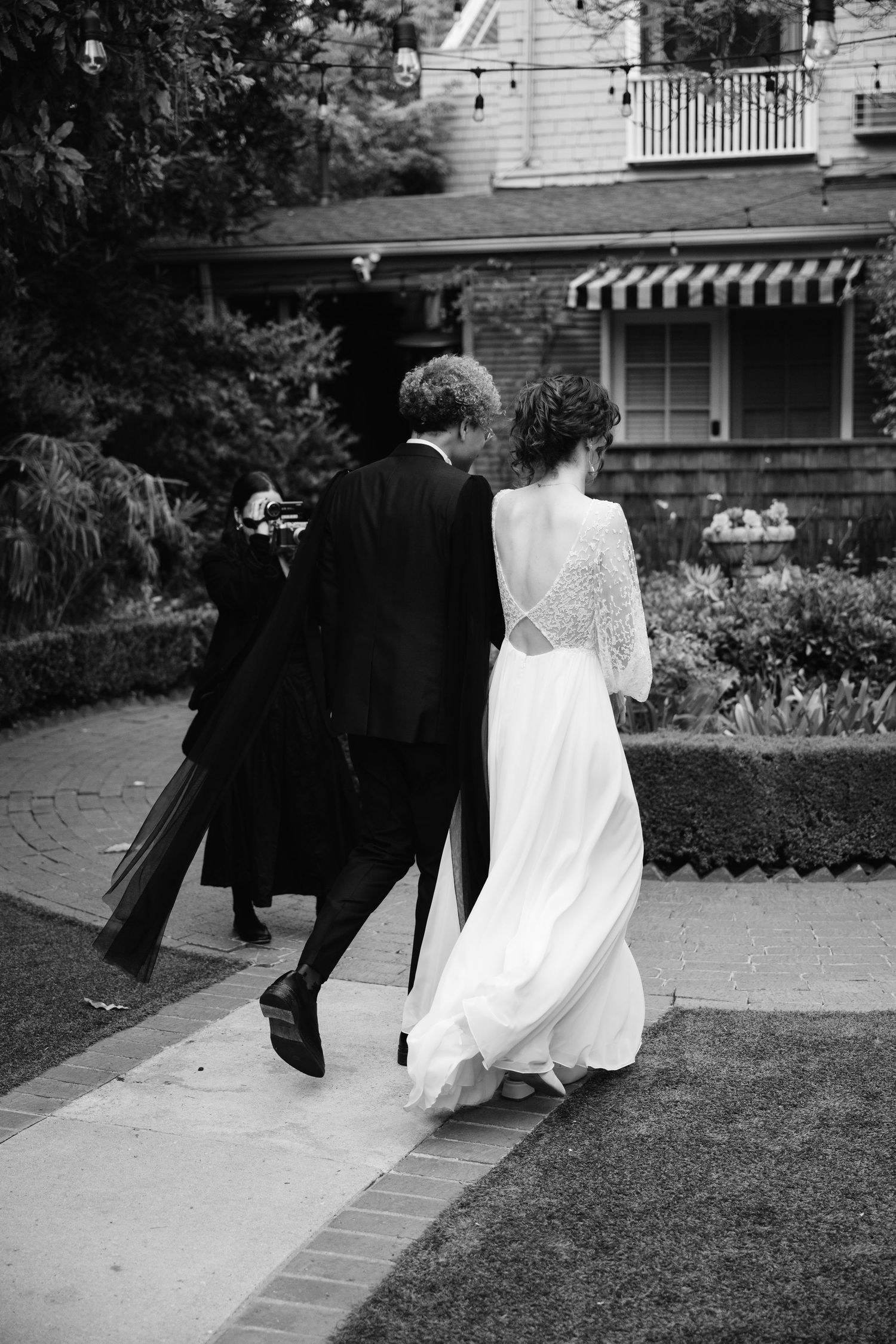 Natural romantic editorial documentary scene of a couple walking a garden path toward a house during a wedding celebration.