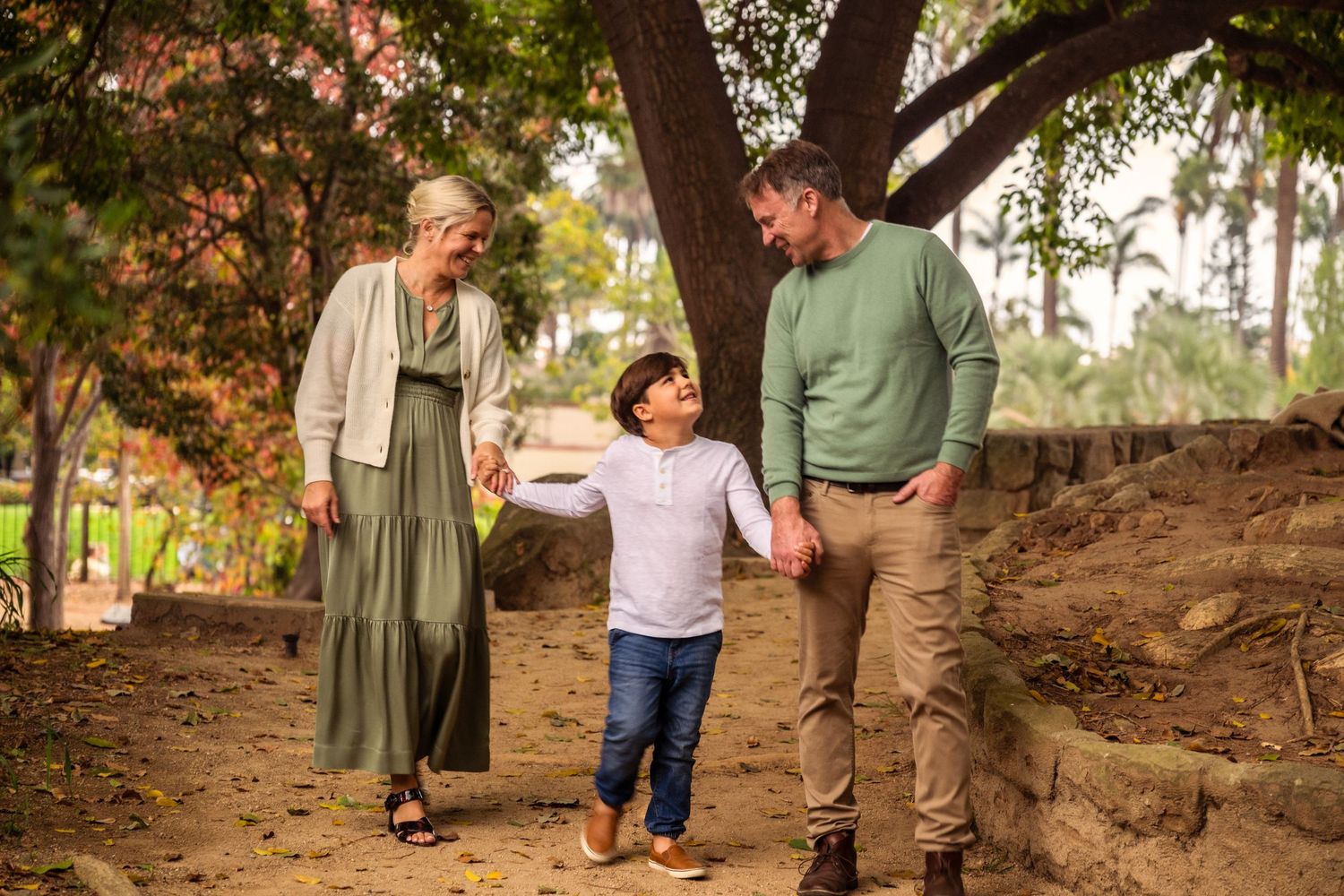 Editorial documentary family photo of two adults walking with a child between them on a sunlit garden path surrounded by trees and greenery.