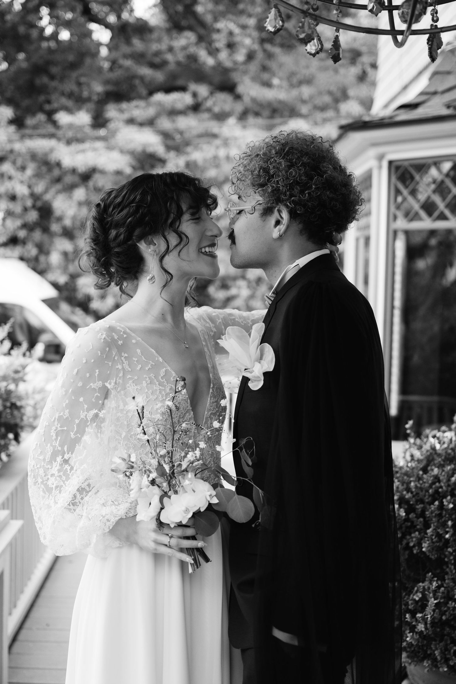 Natural editorial documentary wedding moment of a couple standing close on a porch, holding flowers and sharing an intimate glance.