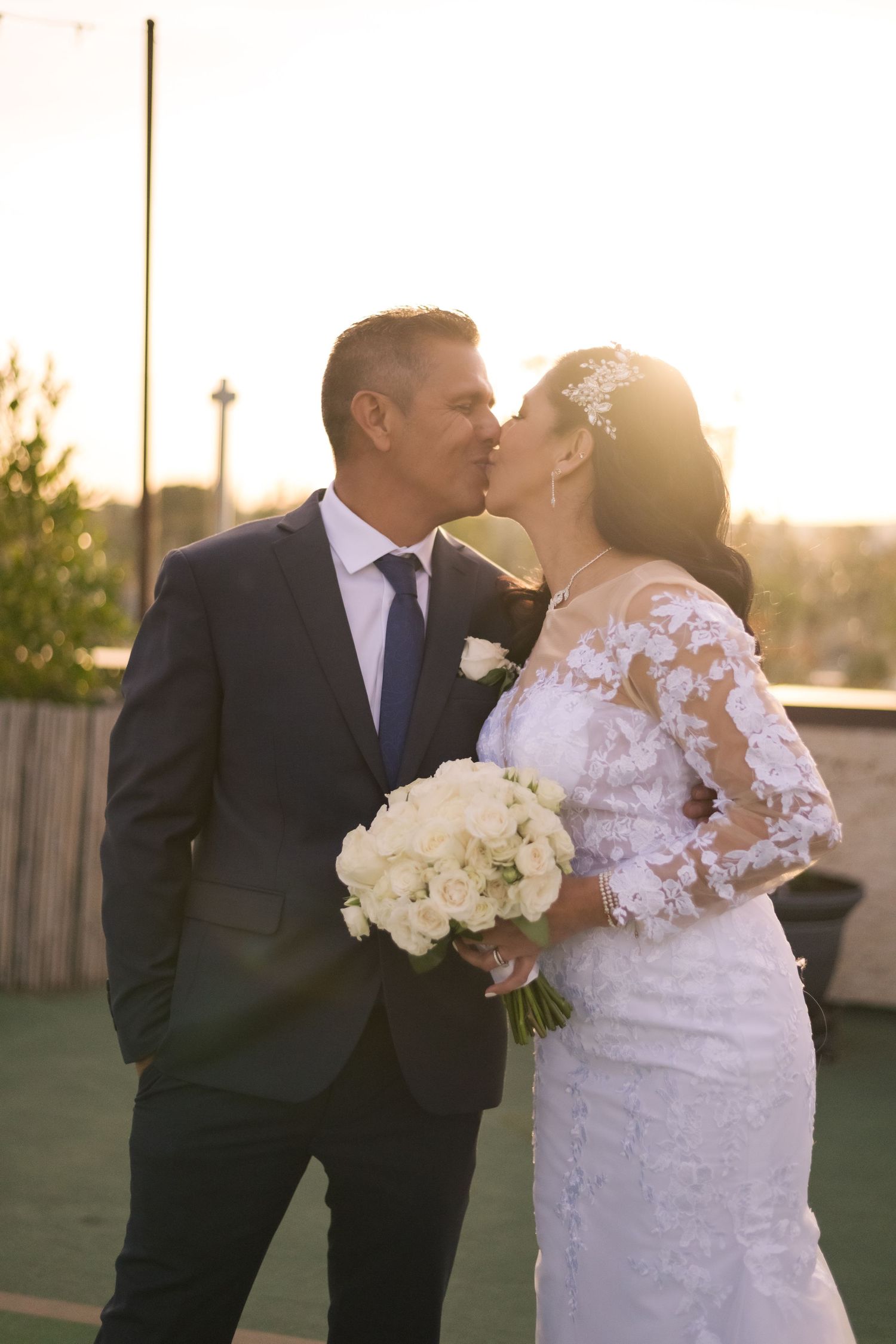 Natural editorial documentary wedding moment of a couple sharing a sunset kiss, bouquet in hand, in warm golden light.