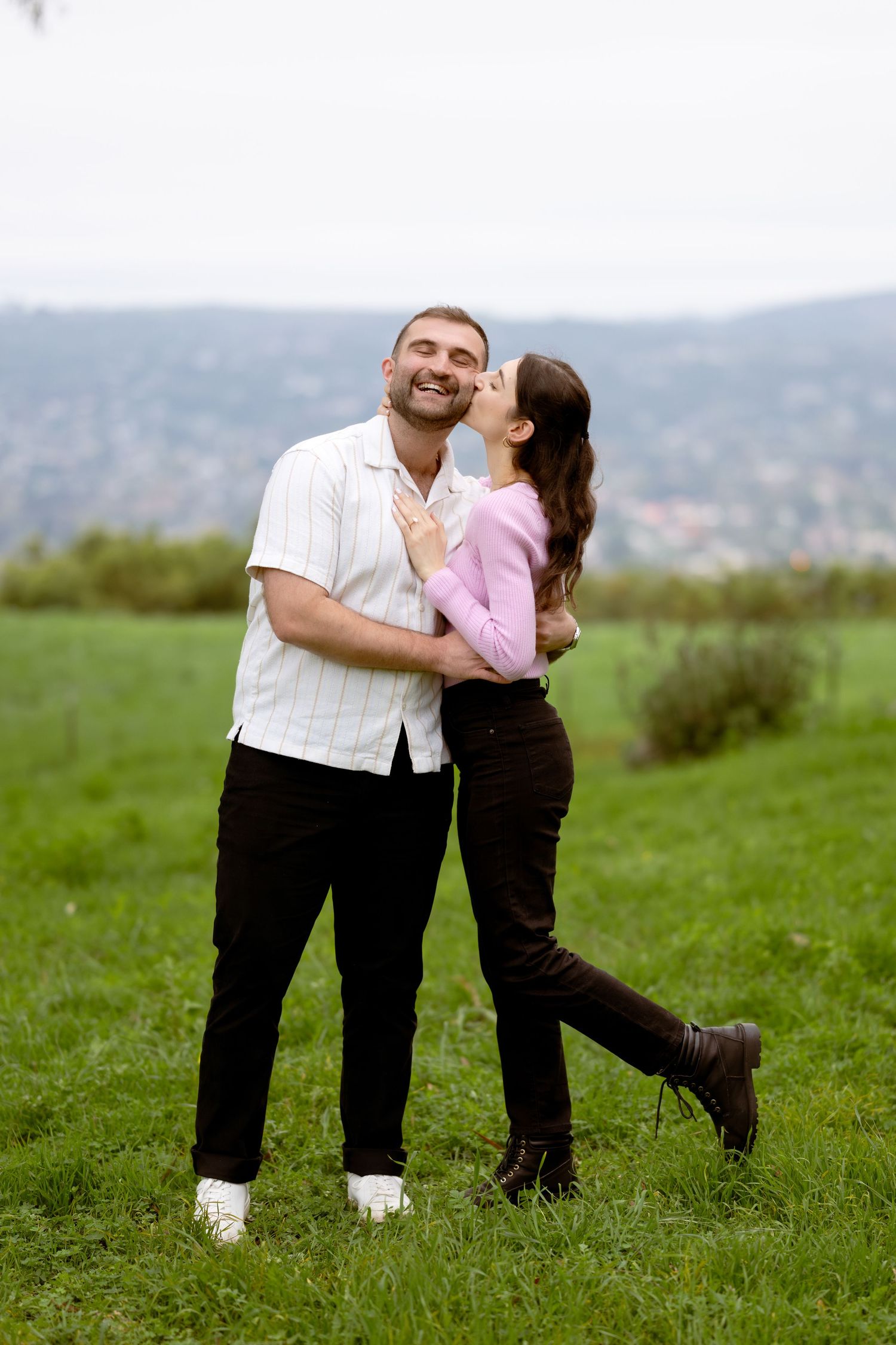 Woman kissing her partner on the cheek in a green field at Franceschi Park Santa Barbara engagement, Herics Photo