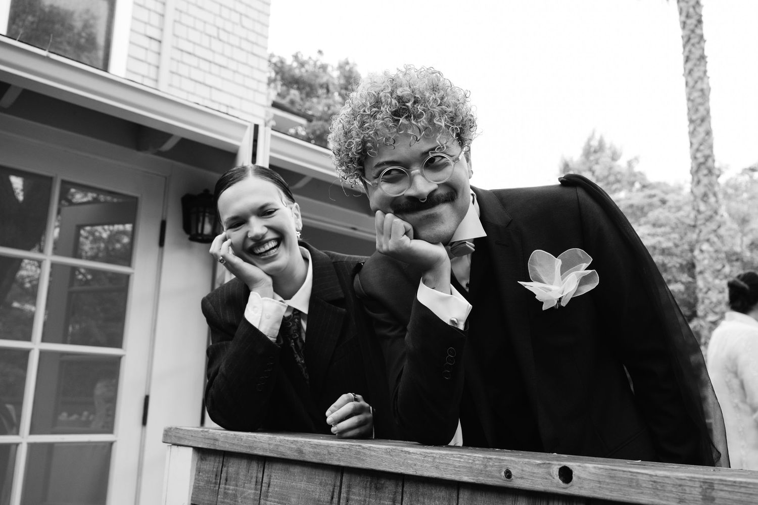 Natural editorial documentary wedding photo of two people in suits posing closely on a railing with soft outdoor light in black and white.