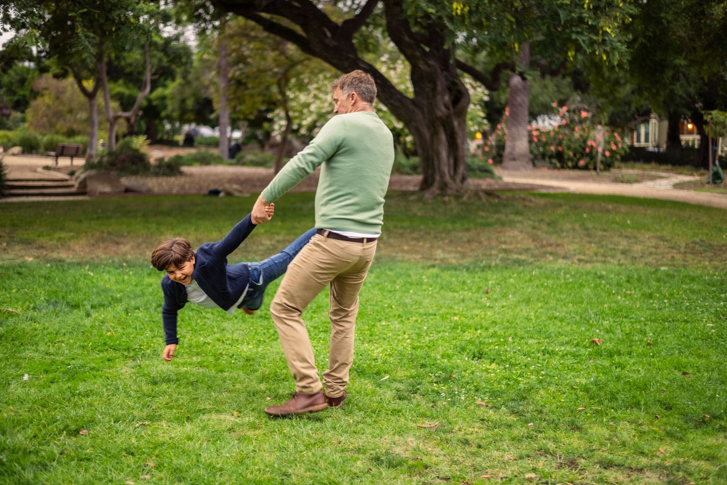 Editorial documentary photo of an adult spinning a child in a sunny park, capturing playful movement and natural outdoor family joy.