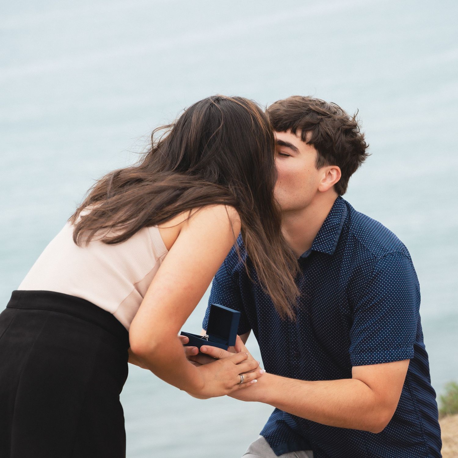 Editorial documentary waterfront proposal moment with one partner kneeling and offering a ring as they kiss in soft natural light.