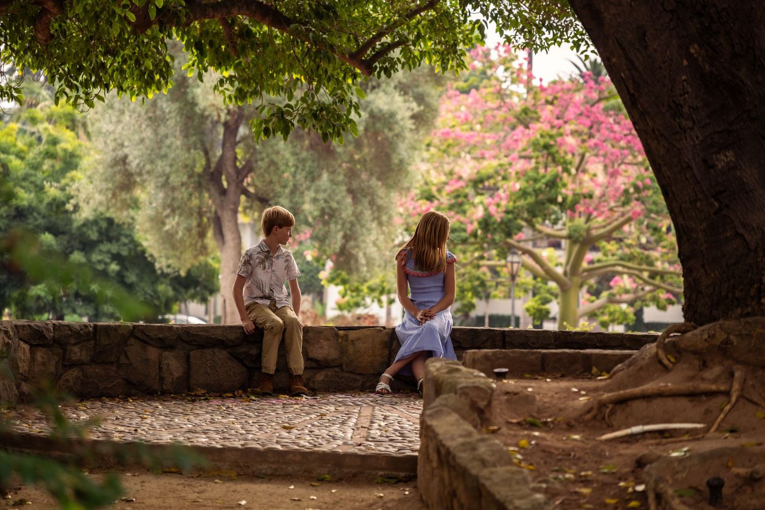 Editorial documentary photo of two kids sitting on a stone wall in a park surrounded by trees, greenery, and soft natural light.