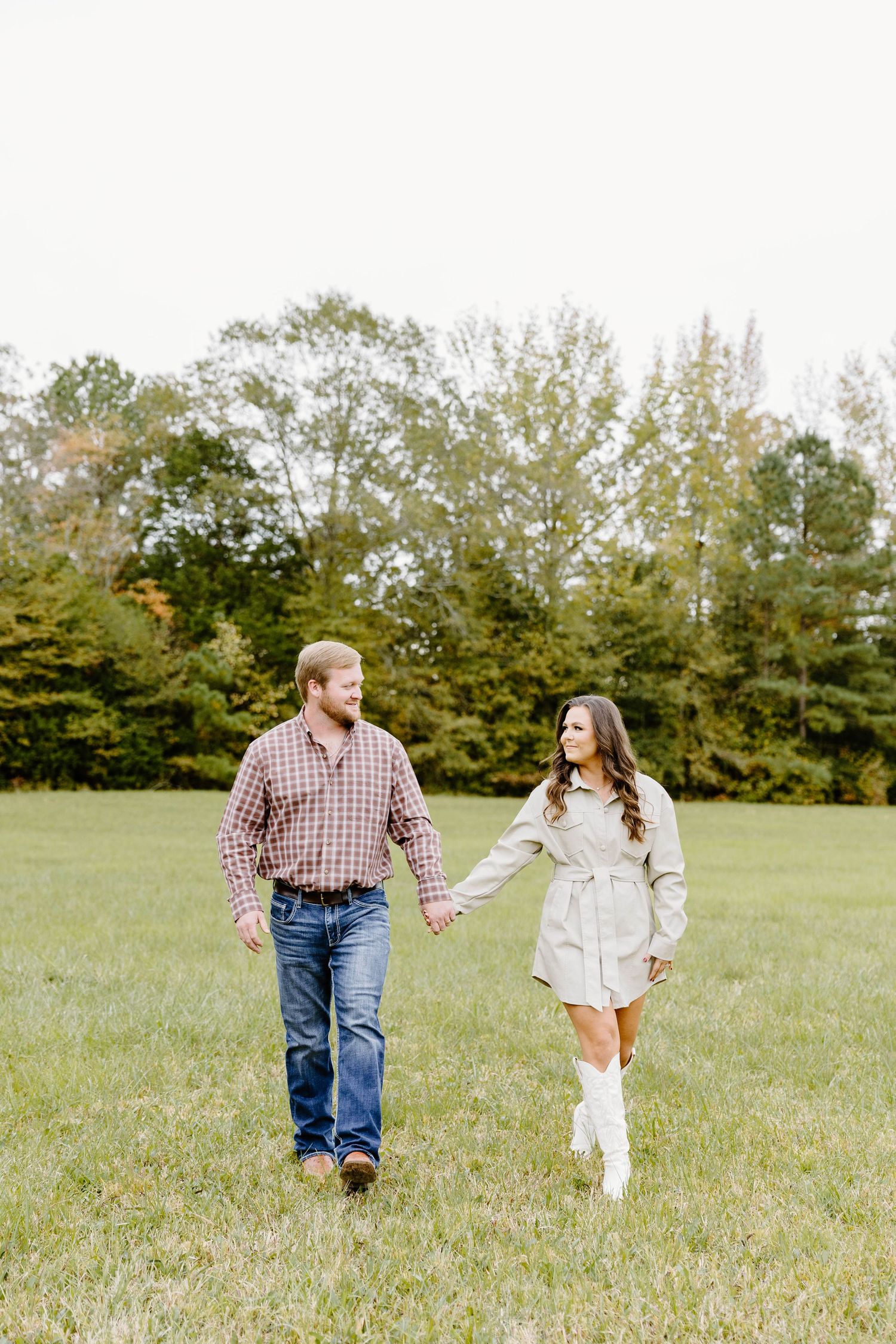 A couple holds hands while walking together through a grassy field on a sunny autumn day.