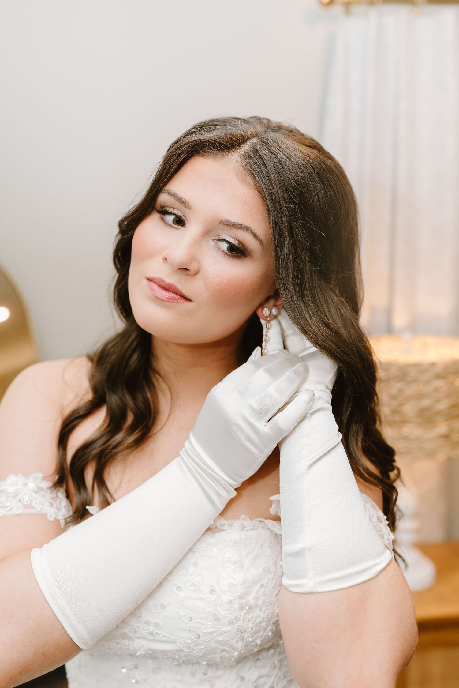 Elegant bridal portrait featuring white satin gloves and delicate jewelry in a softly lit indoor setting.