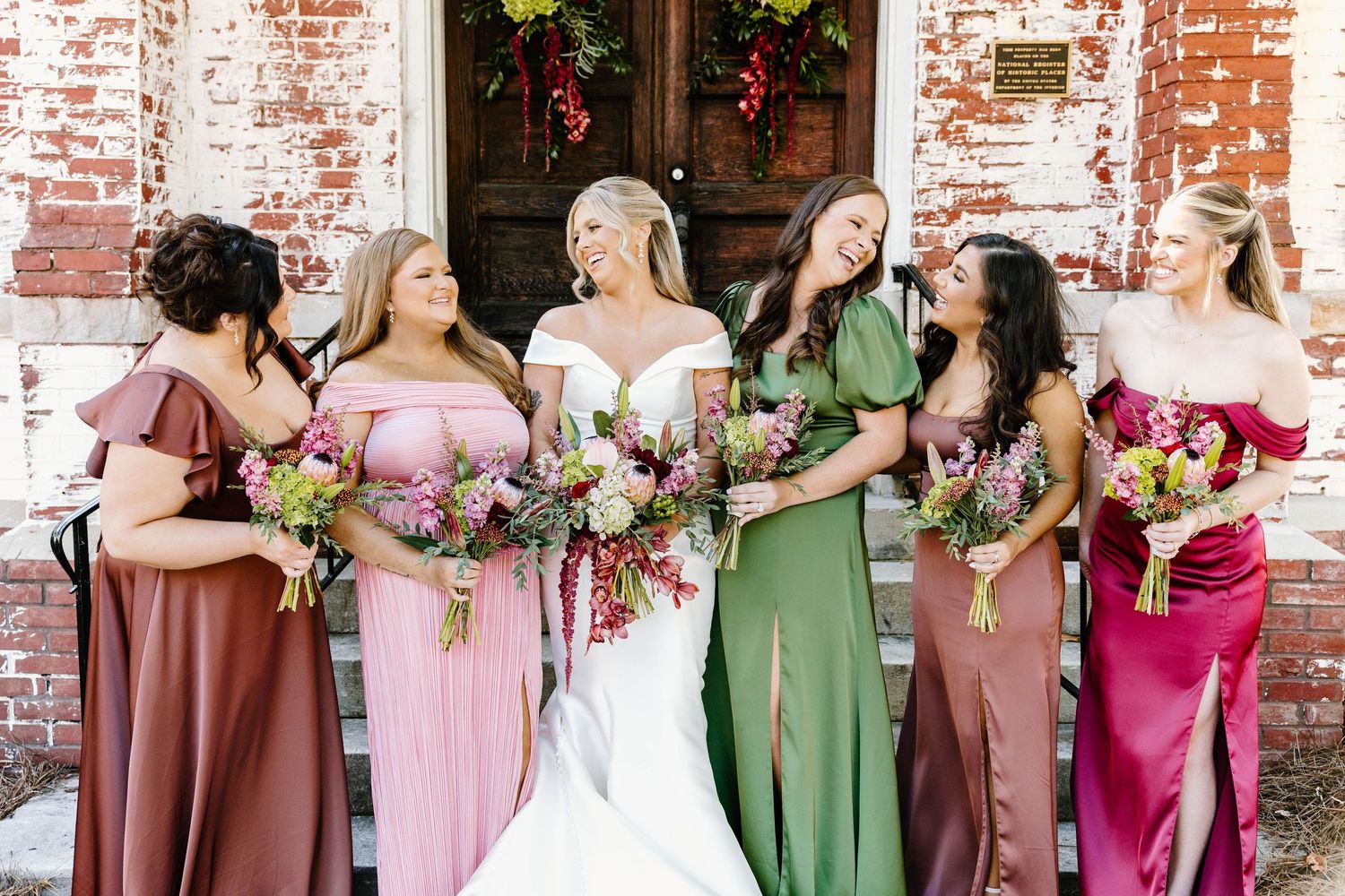 Bridesmaids wearing mix of pink and green dresses pose against rustic brick wall.