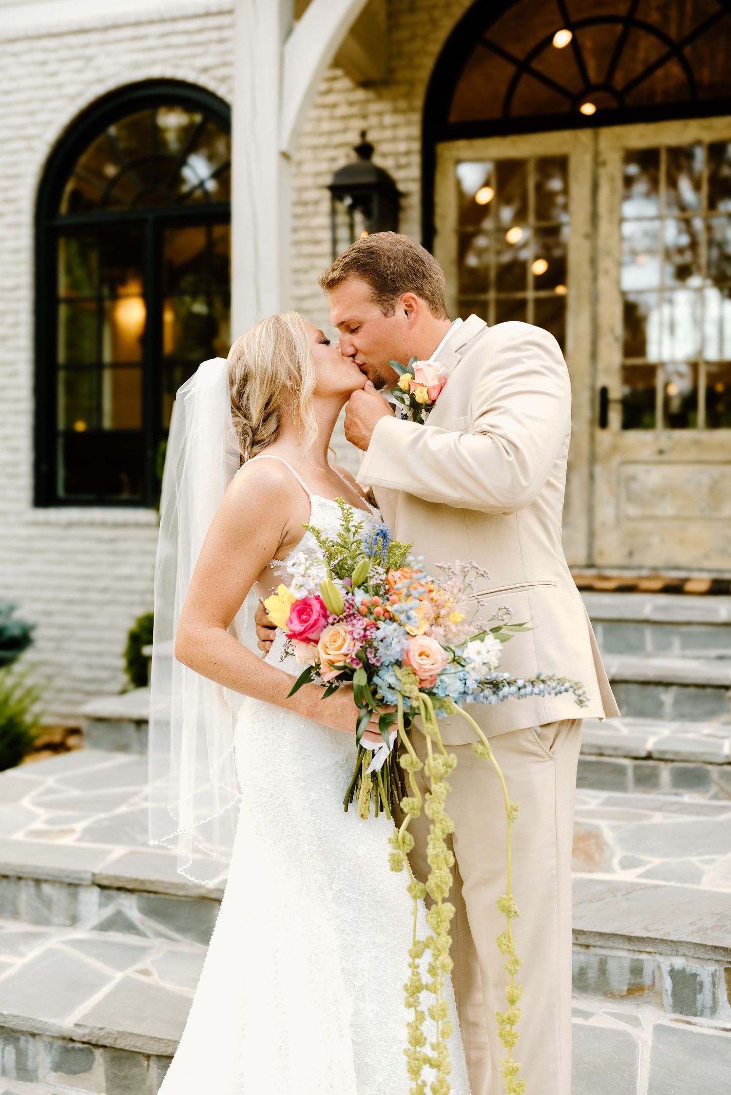 Wedding couple shares romantic moments outside white brick venue with colorful bouquet.